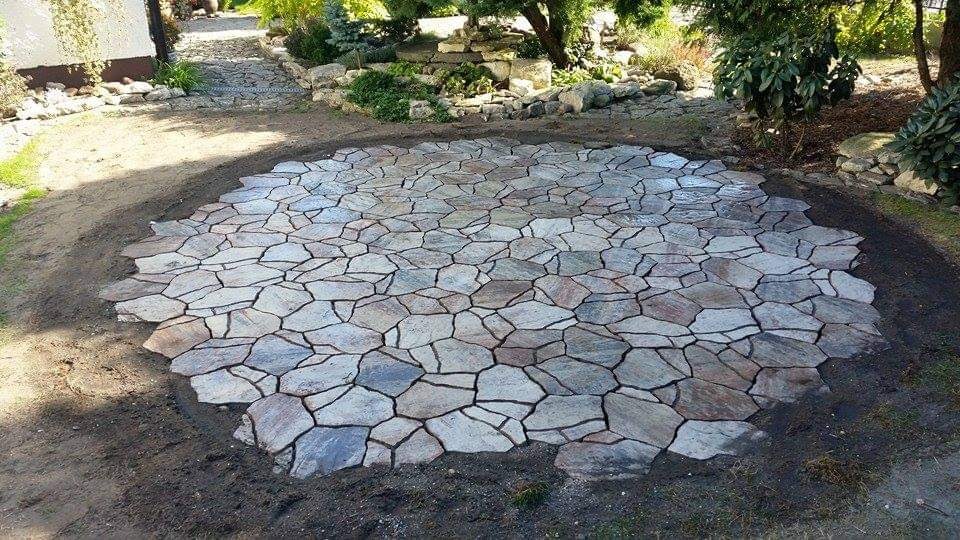 Okrągła powierzchnia wyłożona nieregularną, szarą kostką granitową, otoczona ziemią. Widok na ogród z drzewami i ścieżką z kamienia w tle.