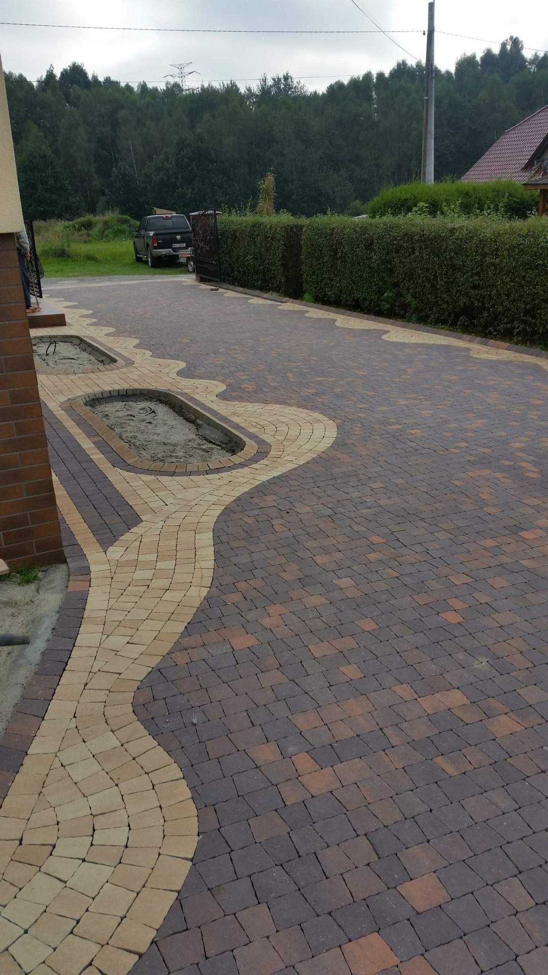 Ukończony podjazd z kostki brukowej w dwóch kolorach, z wyznaczonymi miejscami na zieleń. W tle widoczny samochód i żywopłot.