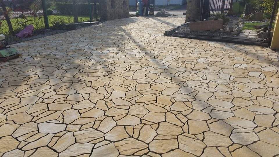 Ukończona nawierzchnia z nieregularnych, beżowych płyt kamiennych, tworząca ścieżkę w ogrodzie. W tle brama wjazdowa i zieleń. Widoczne cienie drzew.