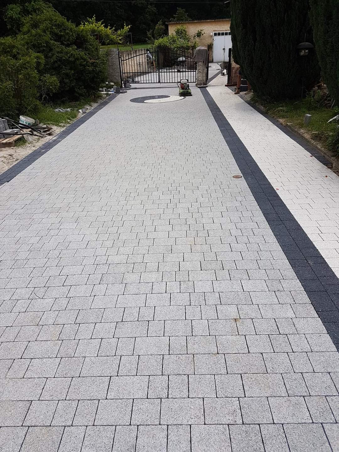 Ułożona jasnoszara kostka brukowa z ciemnymi obrzeżami prowadząca do bramy wjazdowej. W tle budynek i zieleń. Widoczna zagęszczarka w oddali.
