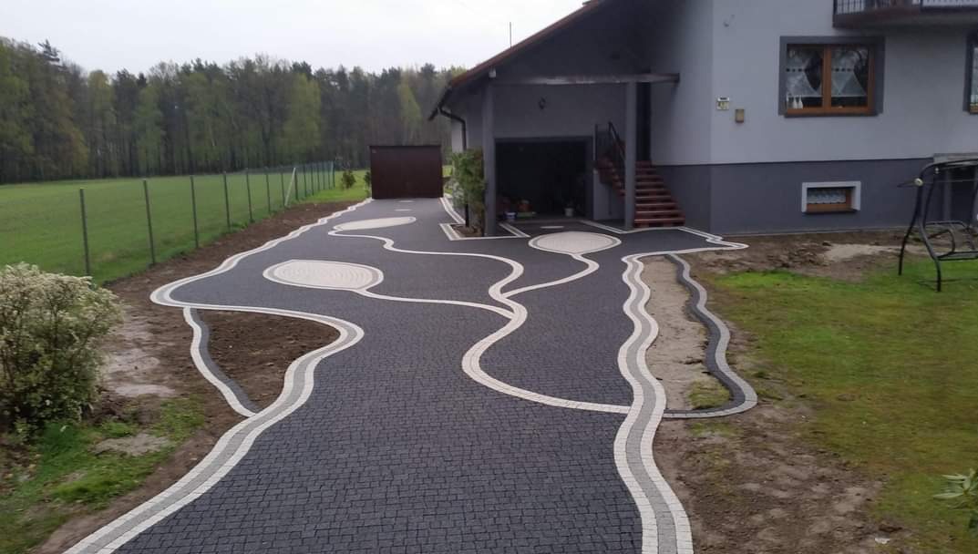 Kreatywny wzór z ciemnej kostki brukowej z jasnymi obwódkami prowadzący do domu z garażem; widok na ogród i las w tle.