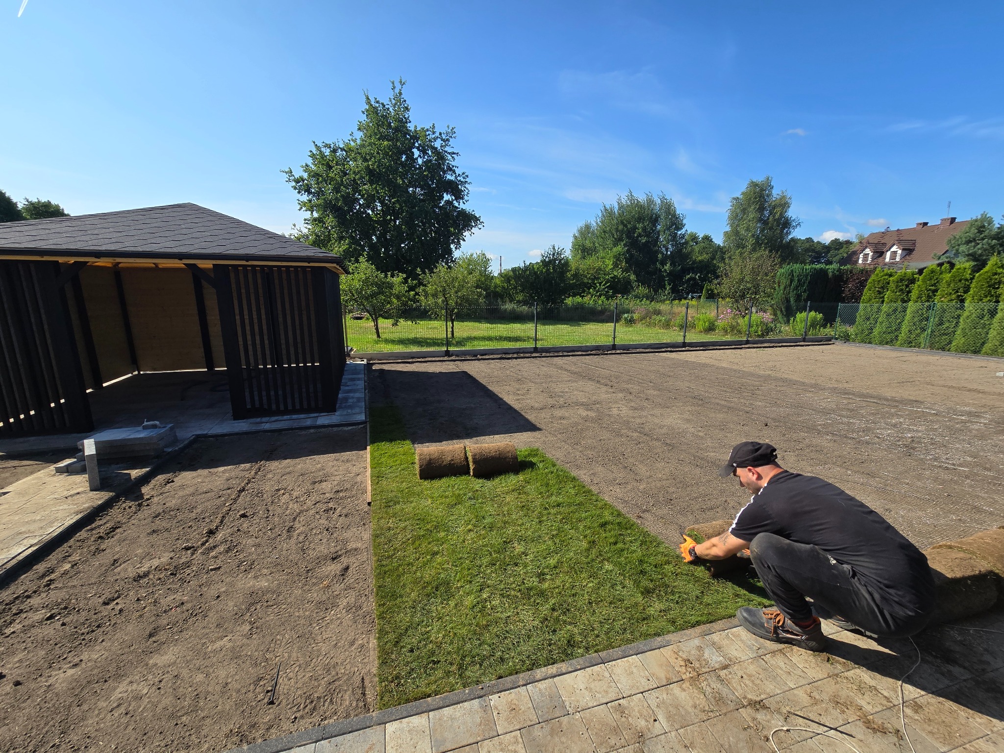 Układanie trawy z rolki na przygotowanym terenie. Widoczny fragment ułożonej trawy, rolki trawy oraz pracownik w trakcie pracy. W tle altana i zieleń.