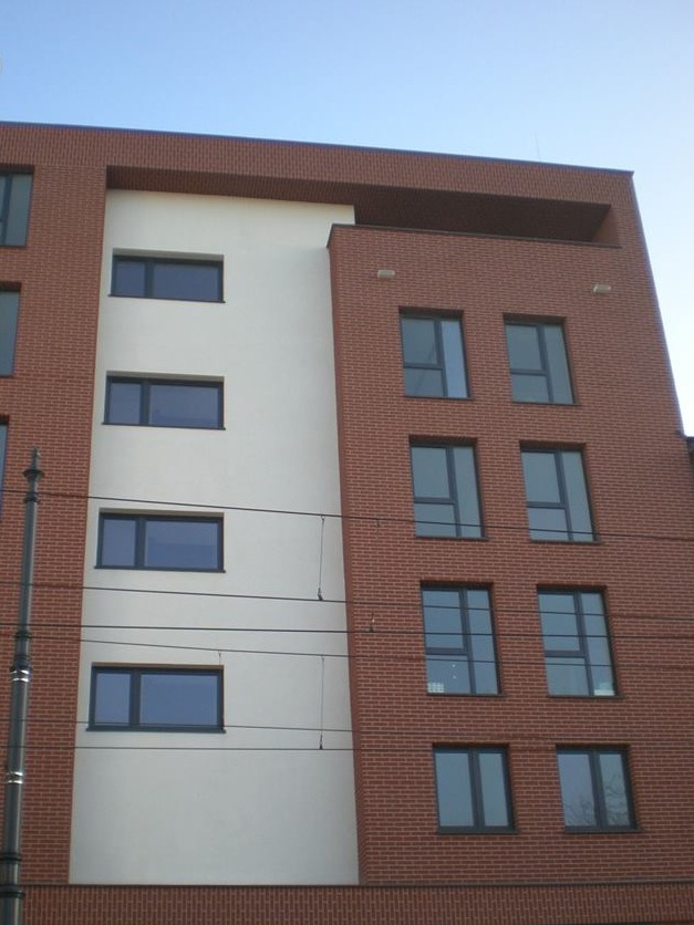 Ceglany budynek z białym fragmentem fasady i ciemnymi oknami. Widok z dołu na nowoczesną architekturę miejską w słoneczny dzień, z widocznymi przewodami elektrycznymi.