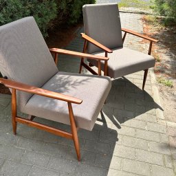 ZURFAM Wojciech Żurawiński - Dwa szare fotele z drewnianymi podłokietnikami na tle kostki brukowej i zieleni. Fotele tapicerowane, w stylu vintage, ustawione na zewnątrz w słoneczny dzień.