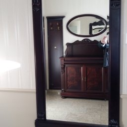 ZURFAM Wojciech Żurawiński - Eleganckie lustro w ciemnej, zdobionej ramie, odbijające klasyczną komodę i szafę w przedpokoju. Widoczne detale wykończenia i odbicie osoby.