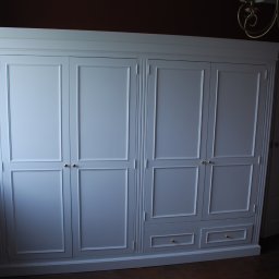 ZURFAM Wojciech Żurawiński - Elegancka, biała szafa czterodrzwiowa z szufladami w dolnej części, z dekoracyjnymi listwami i złotymi uchwytami, ustawiona w pokoju z ciemną ścianą i widocznym żyrandolem.