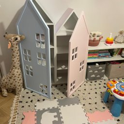 ZURFAM Wojciech Żurawiński - Dwie szafki w kształcie domków, jedna błękitna, druga różowa, z wyciętymi okienkami, stoją w pokoju dziecięcym obok zabawek i regału.