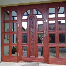 ZURFAM Wojciech Żurawiński - Stylowe, mahoniowe drzwi wejściowe z szybami, zdobione rzeźbieniem i uchwytem w kształcie lwa, odbijające otoczenie. Widok z bliska.