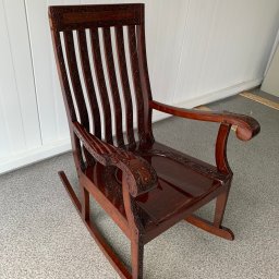 ZURFAM Wojciech Żurawiński - Ręcznie rzeźbiony, mahoniowy fotel bujany z wysokim oparciem i ozdobnymi podłokietnikami na tle białej boazerii i szarej podłogi.