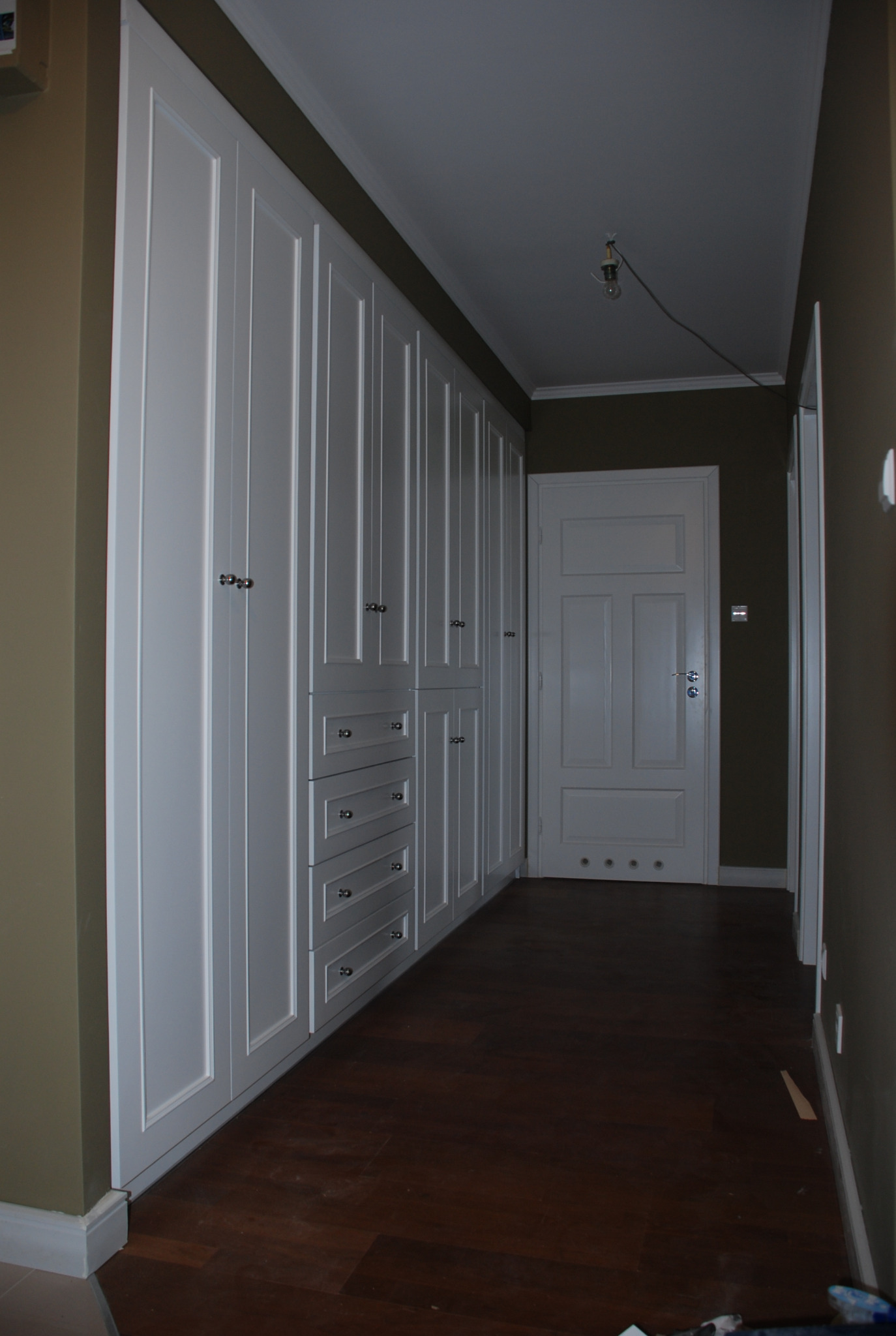 Długa, biała szafa wnękowa z frezowanymi drzwiami i szufladami, z ciemną podłogą i oświetleniem sufitowym w tle, widziana w korytarzu.
