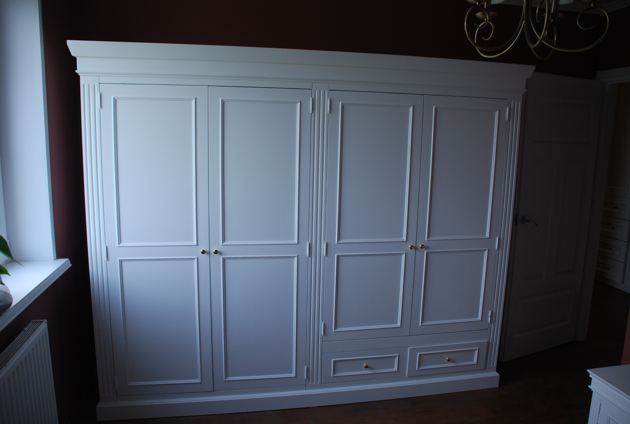 Elegancka, biała szafa czterodrzwiowa z szufladami w dolnej części, z dekoracyjnymi listwami i złotymi uchwytami, ustawiona w pokoju z ciemną ścianą i widocznym żyrandolem.