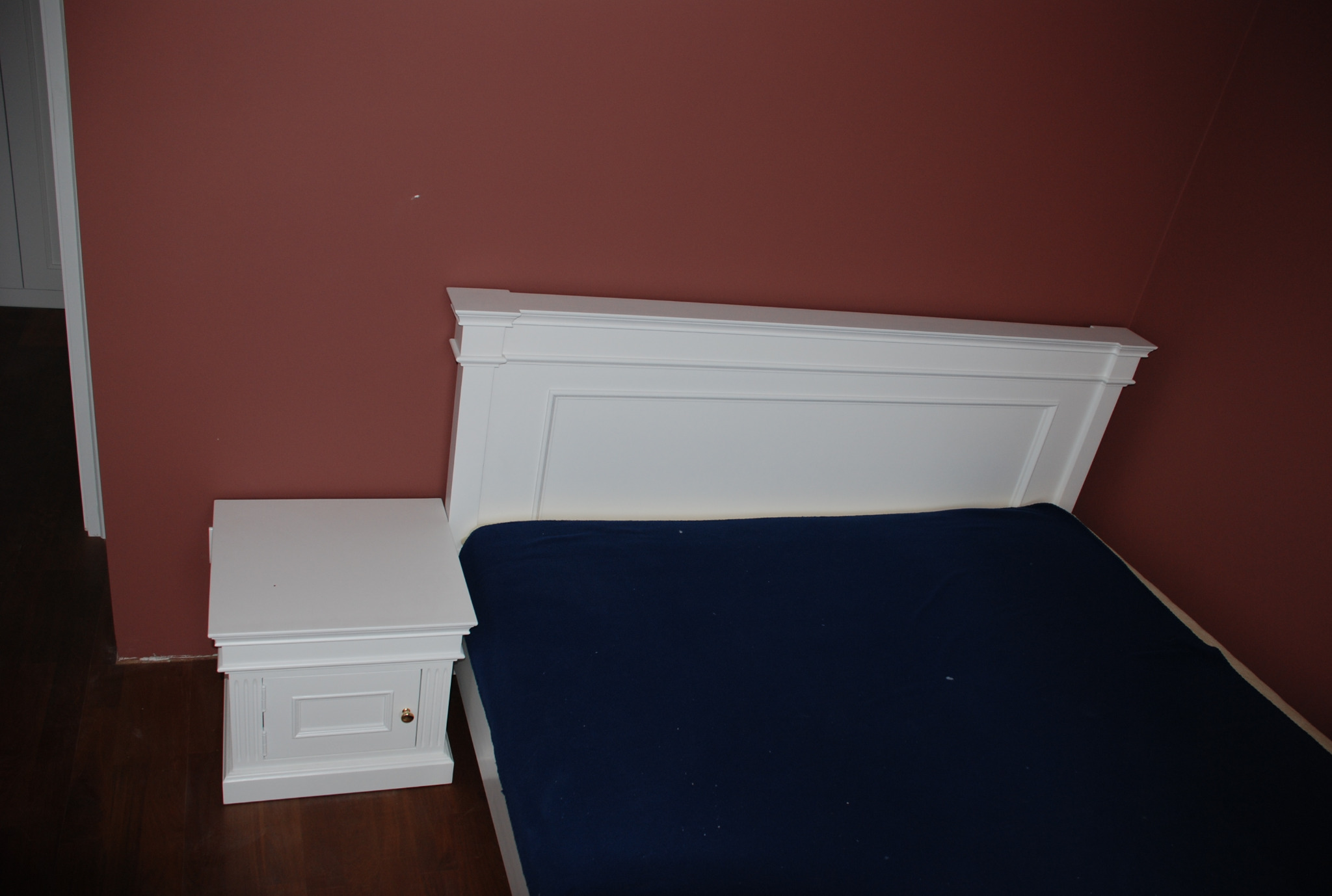 Białe łóżko z szafką nocną w pokoju z bordową ścianą w Pułtusku. Klasyczny styl, widoczny detal frezowania na szafce i wezgłowiu.