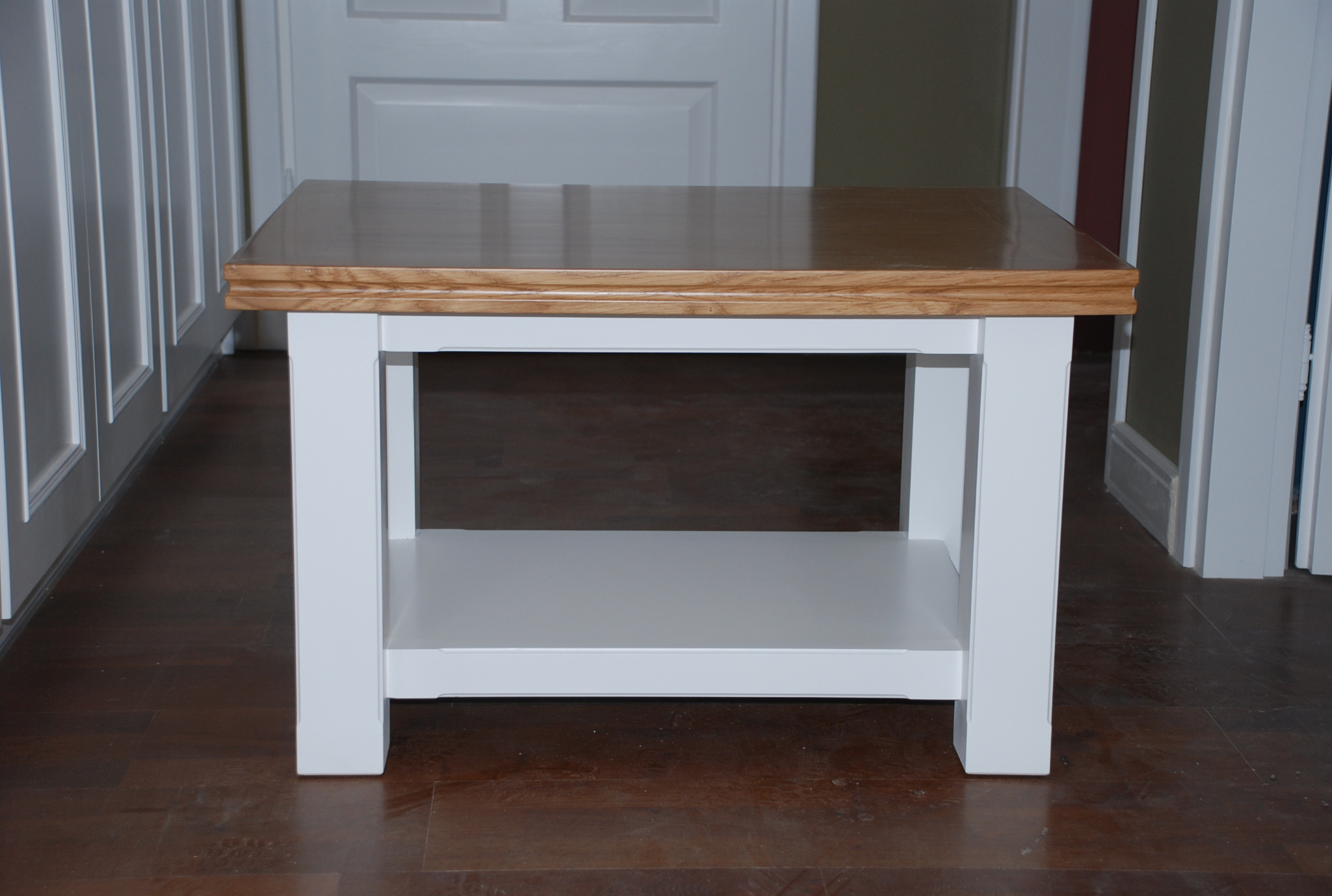 Drewniany stolik kawowy z blatem w odcieniach brązu i białymi nogami, ustawiony na ciemnej podłodze, na tle białych drzwi.