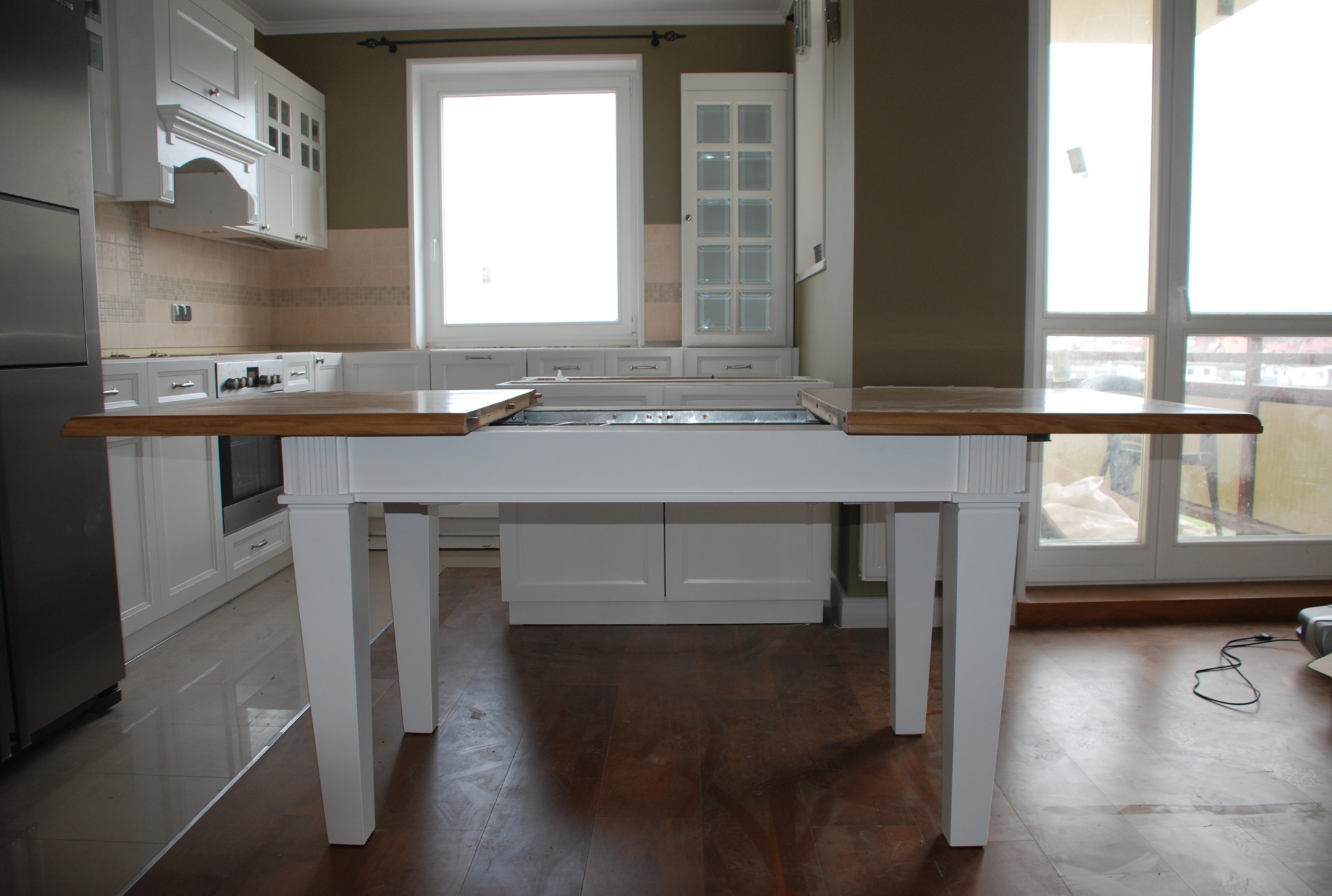 Rozkładany stół z drewnianym blatem i białymi nogami w kuchni z zabudową na wymiar. Widoczny mechanizm rozkładania i fragmenty szafek kuchennych.