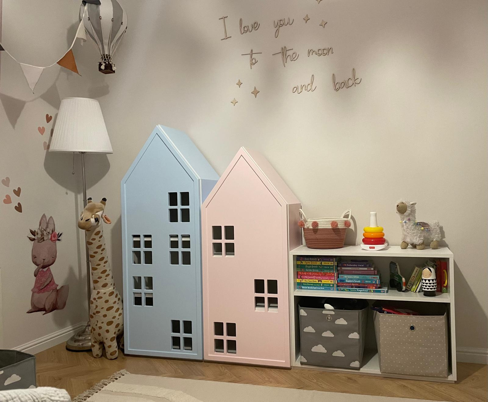 Pokój dziecięcy: pastelowe szafki w kształcie domków (różowa i błękitna) stoją obok regału z zabawkami. Na ścianie dekoracje: balon, serca, napis 'I love you to the moon and back'.