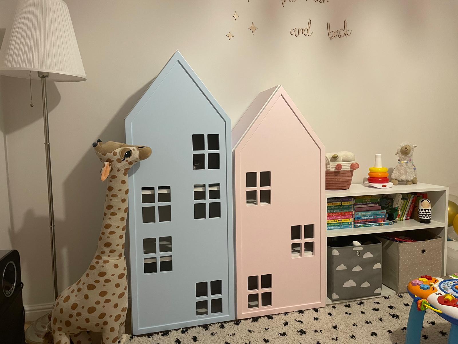 Dwa domki-półki w pastelowych kolorach (różowy i błękitny) w pokoju dziecięcym, obok lampa i pluszowa żyrafa. Oryginalne meble na książki i zabawki.