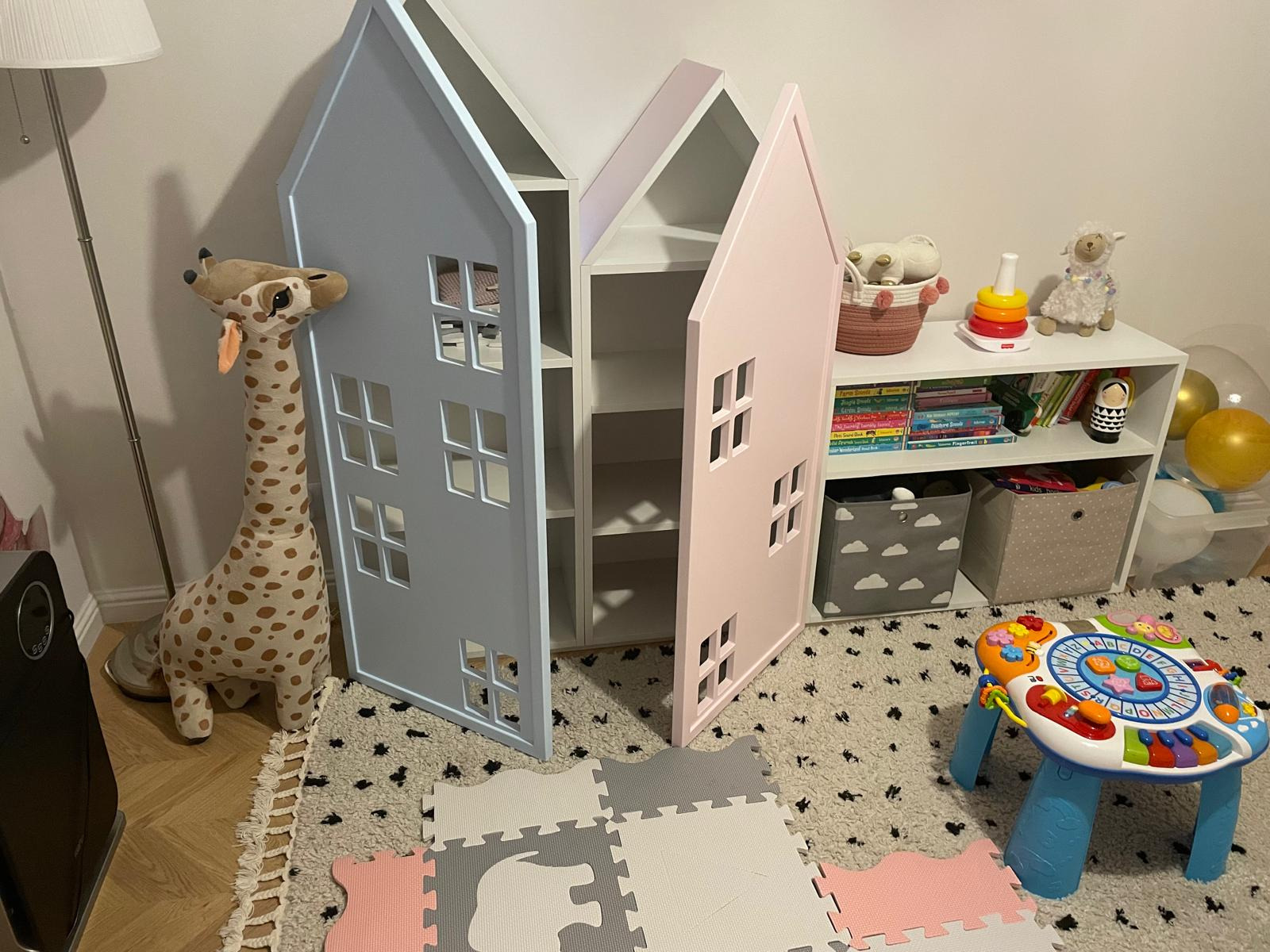 Dwie szafki w kształcie domków, jedna błękitna, druga różowa, z wyciętymi okienkami, stoją w pokoju dziecięcym obok zabawek i regału.