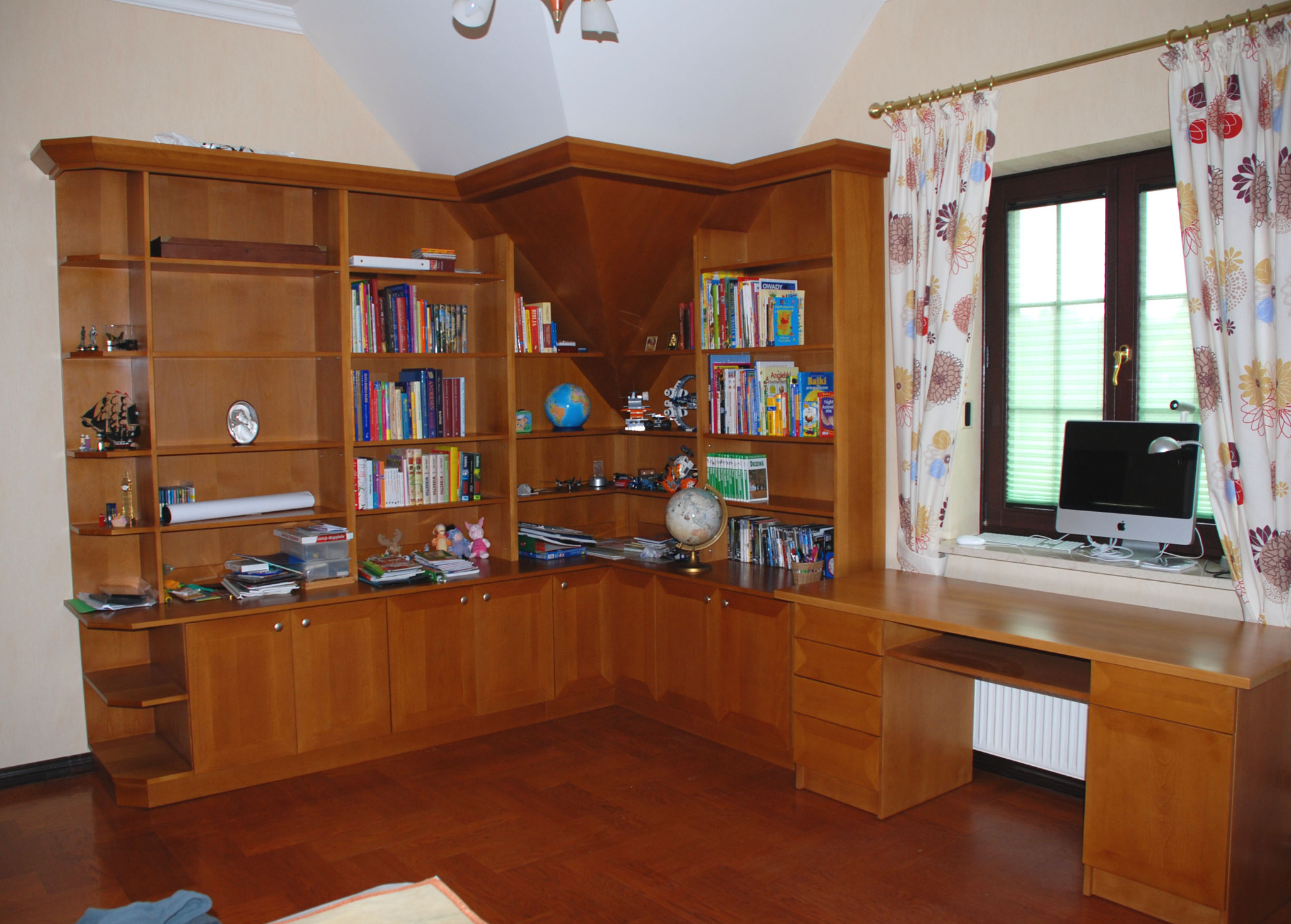 Drewniana zabudowa biblioteczna z biurkiem pod oknem, wypełniona książkami i dekoracjami. Wyjątkowa konstrukcja pod sufitem.