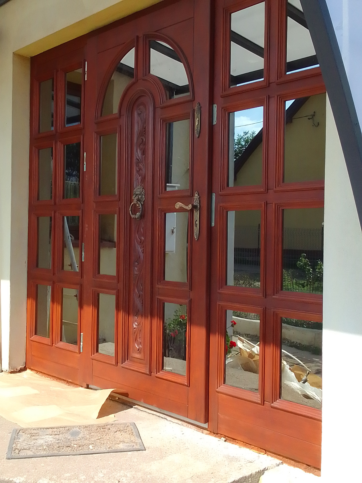 Dwuskrzydłowe, przeszklone, drewniane drzwi wejściowe z dekoracyjnym okuciem i wzorem. Widoczne refleksy otoczenia w szybach.