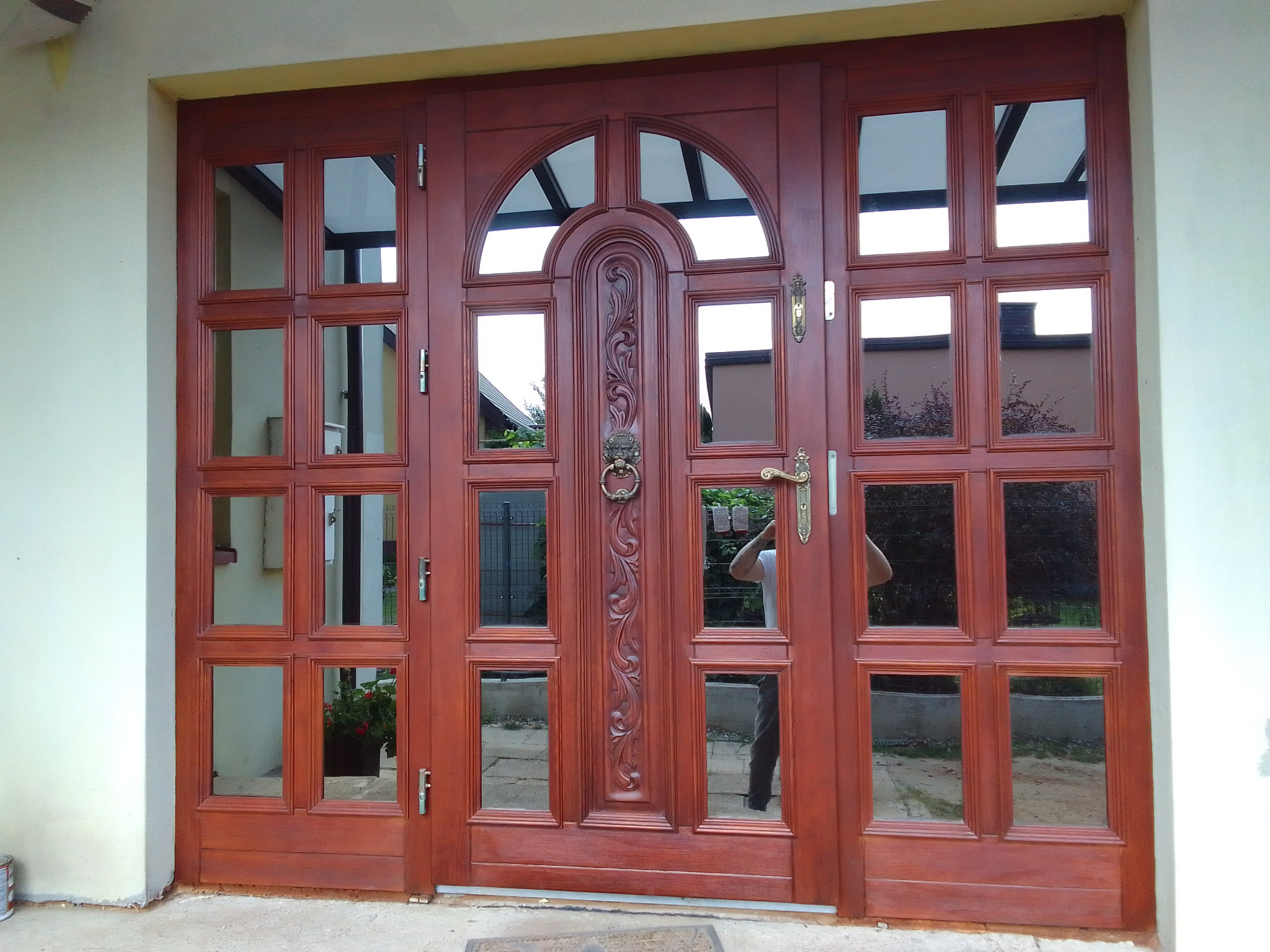 Stylowe, mahoniowe drzwi wejściowe z szybami, zdobione rzeźbieniem i uchwytem w kształcie lwa, odbijające otoczenie. Widok z bliska.