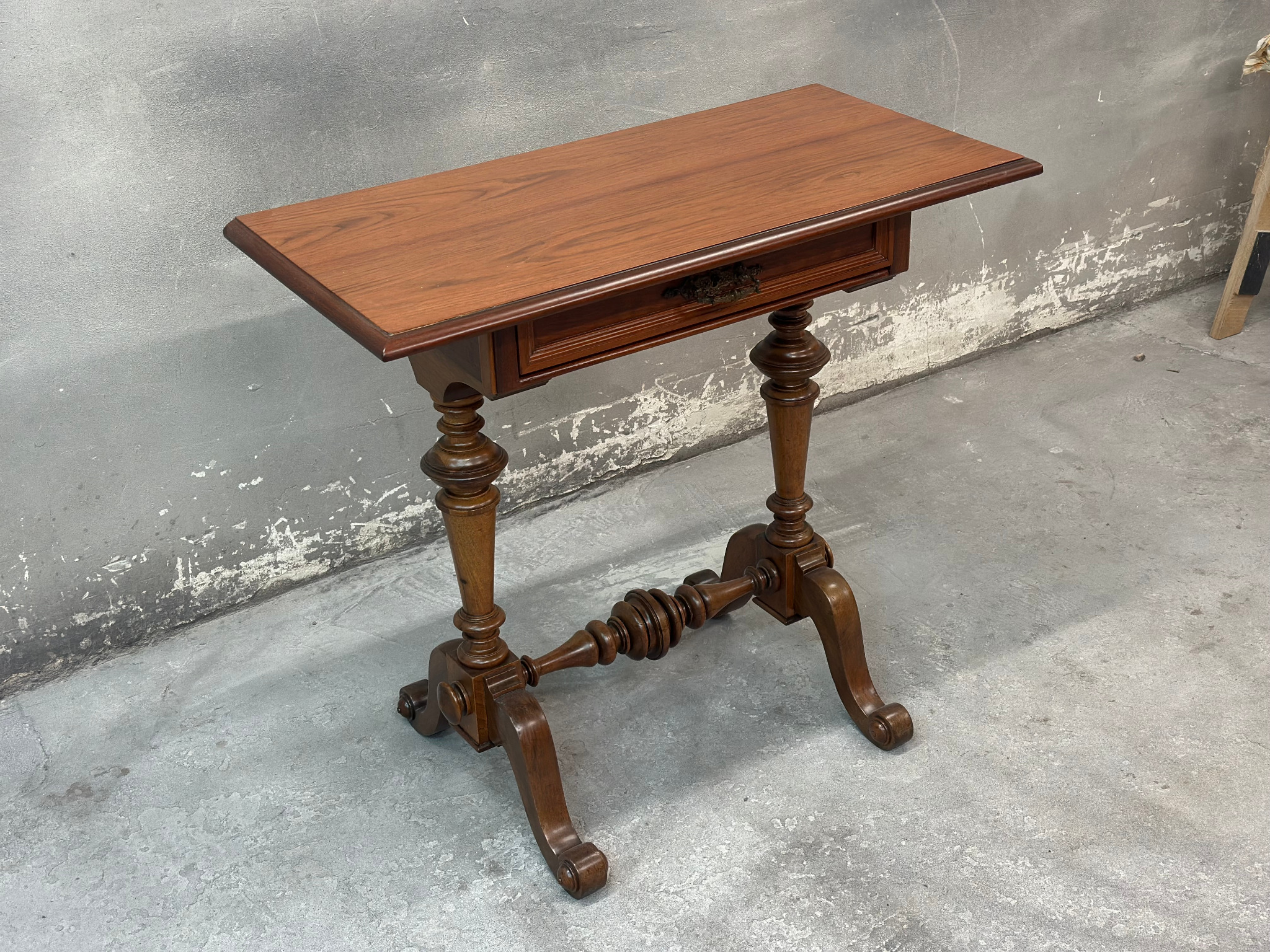 Antyczny stolik z szufladą, wykonany z ciemnego drewna, z rzeźbionymi nogami, na tle szarej ściany i podłogi. Widoczne detale zdobień.