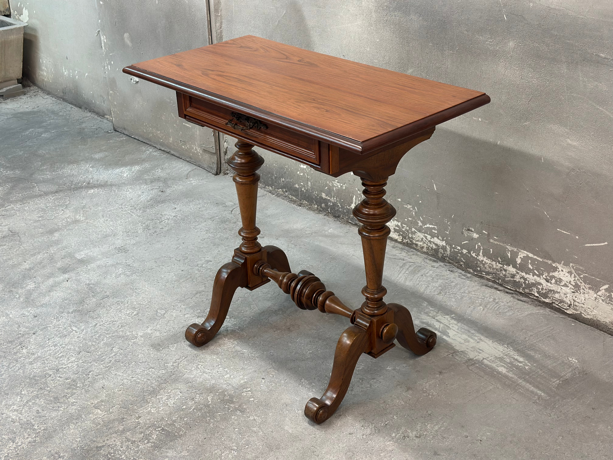 Antyczny stolik z drewna o toczonych nogach i szufladą, na tle surowej ściany betonowej, ukazujący kunszt stolarski i dbałość o detale.