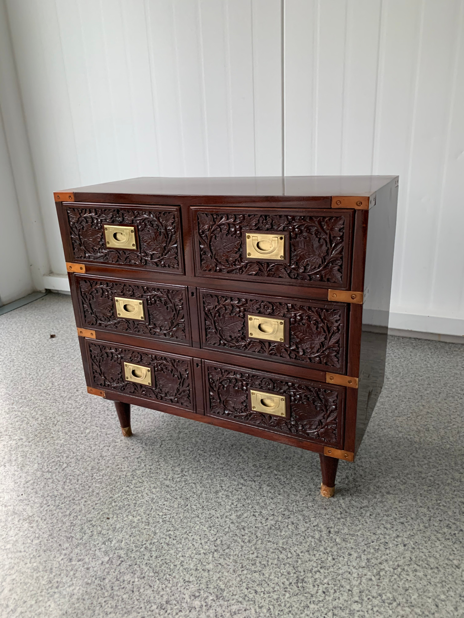 Ręcznie rzeźbiona komoda z ciemnego drewna z mosiężnymi uchwytami i narożnikami, stojąca na smukłych nóżkach. Dekoracyjny, unikatowy mebel do sypialni lub salonu.