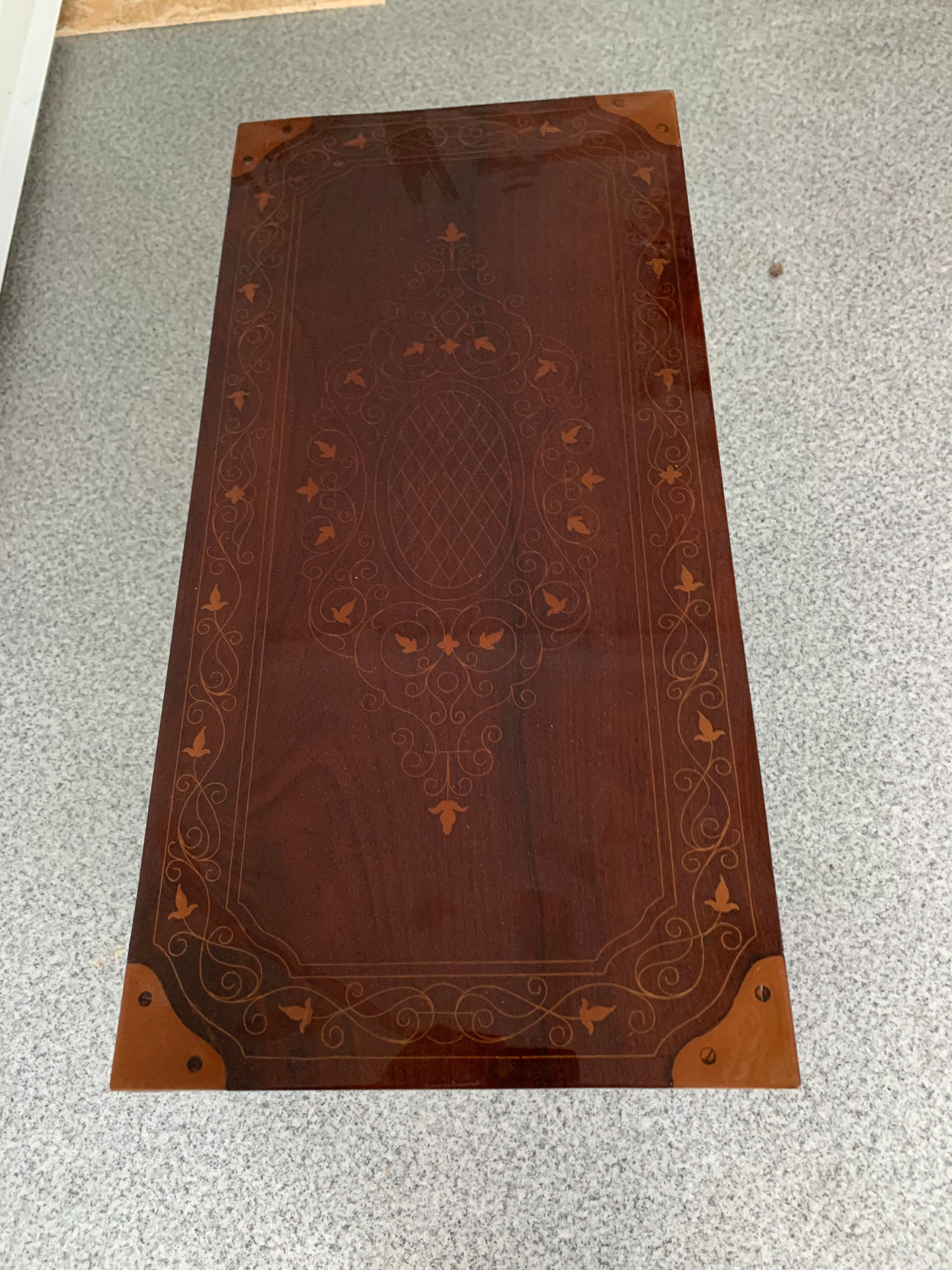 Stylowy, ciemnobrązowy blat stolika kawowego z intarsjami i metalowymi narożnikami, widoczny z góry na tle szarej podłogi. Detale dekoracyjne.