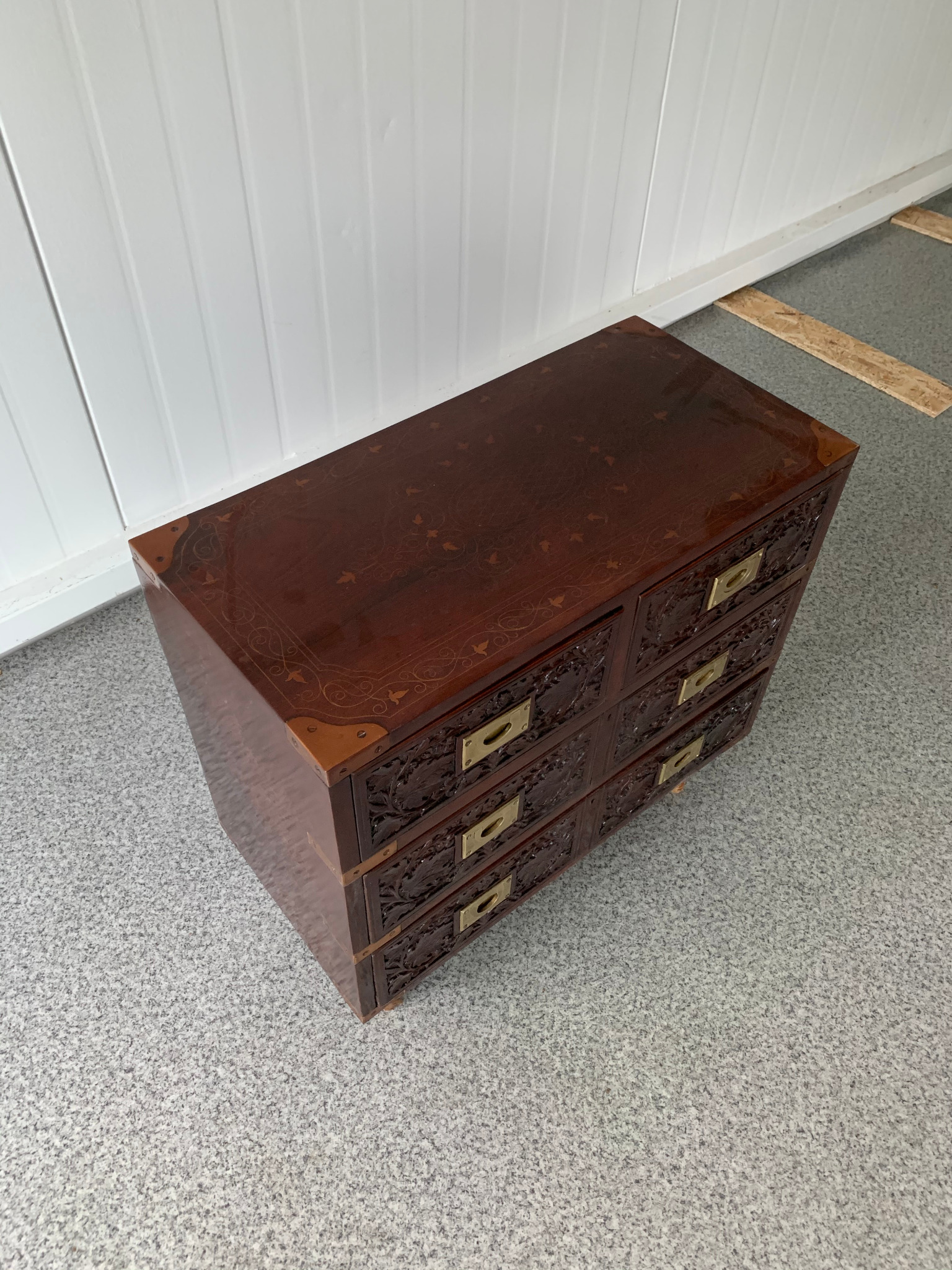 Ręcznie rzeźbiona, mahoniowa komoda z sześcioma szufladami i mosiężnymi uchwytami, wykończona wzorem intarsji na blacie, na tle szarej podłogi.