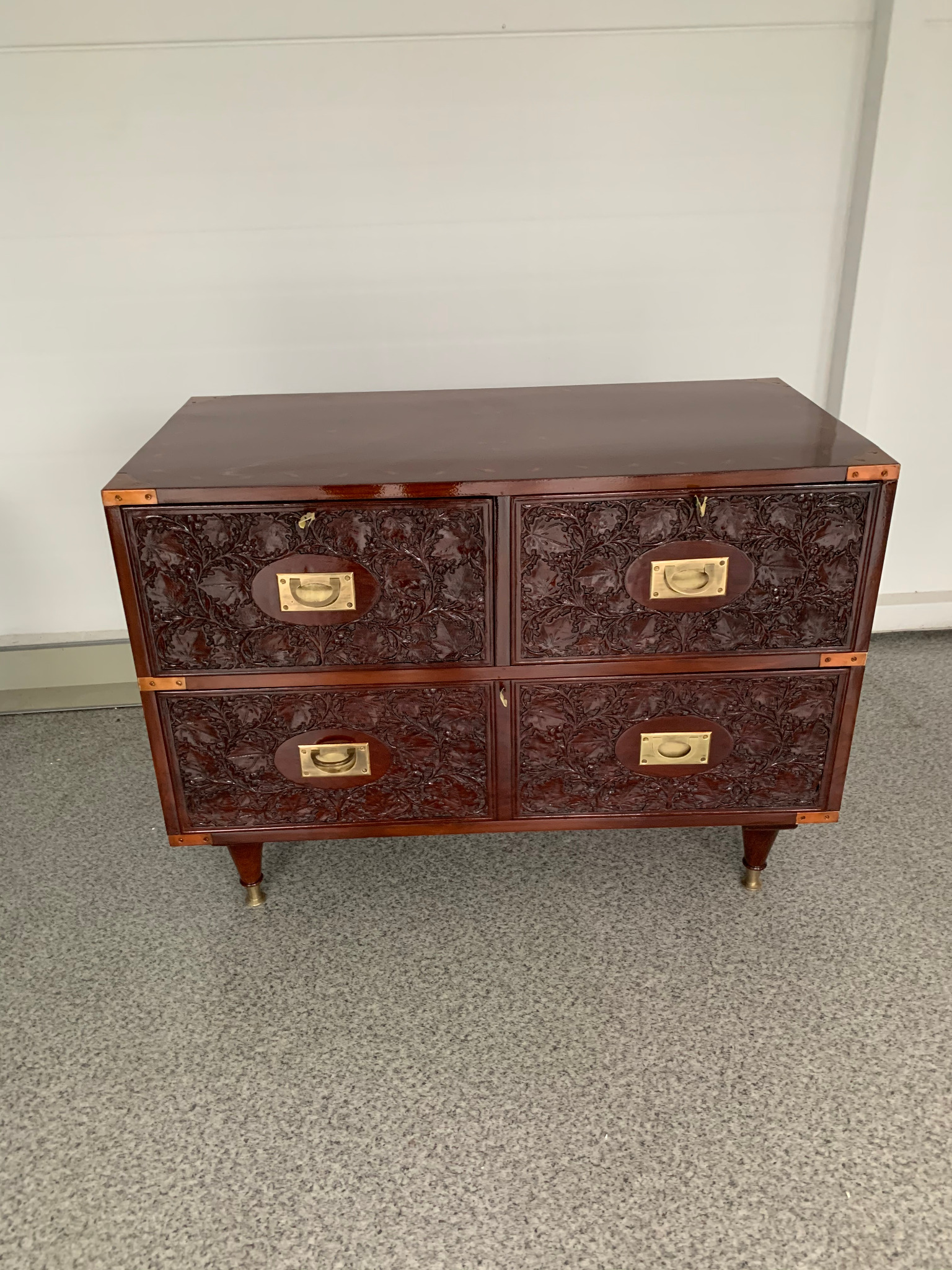 Ciemnobrązowa komoda z czterema szufladami, zdobiona rzeźbieniem i metalowymi okuciami. Stoi na czterech nóżkach z mosiężnymi zakończeniami na tle jasnej ściany.