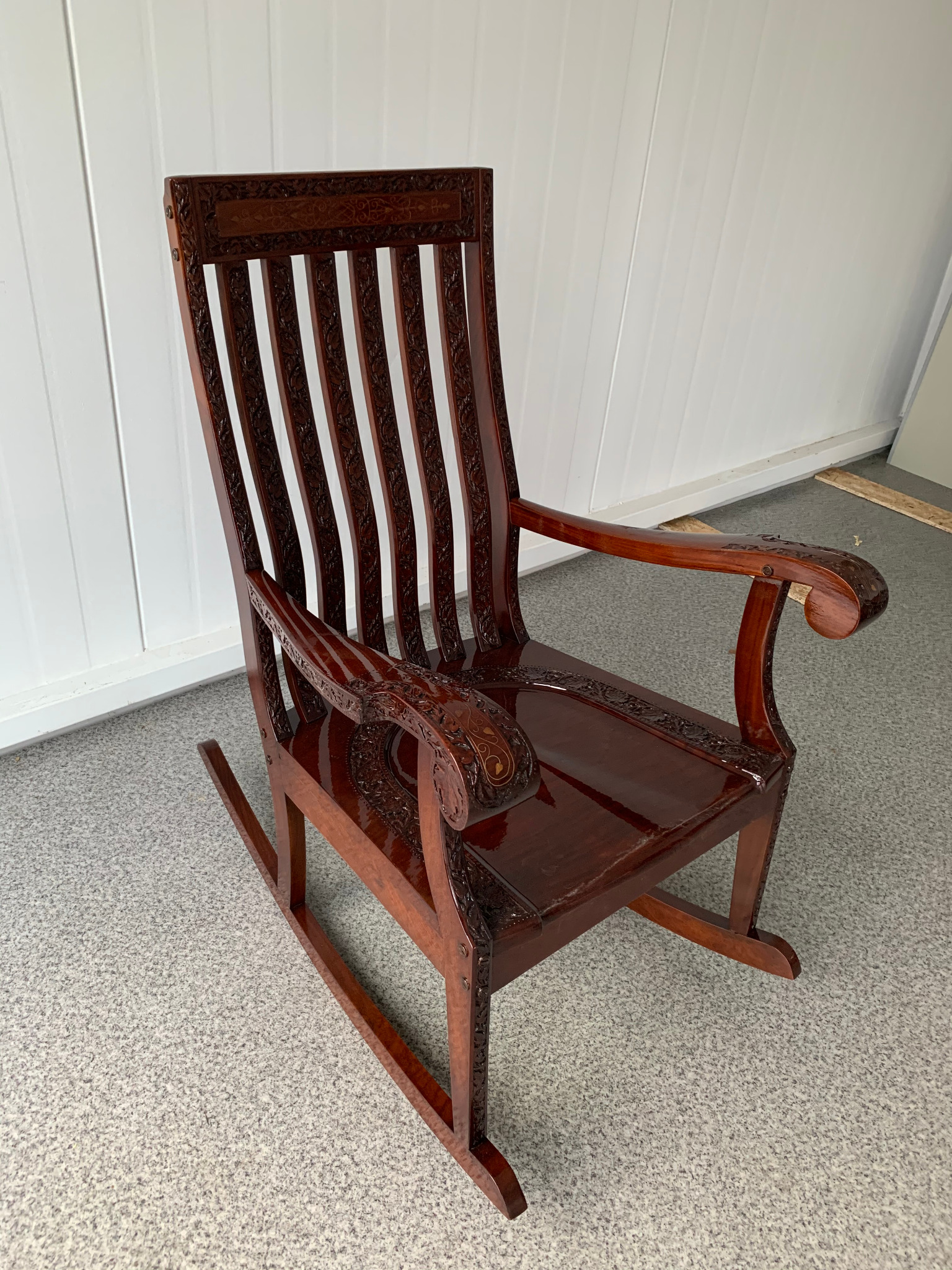 Ręcznie rzeźbiony, mahoniowy fotel bujany z wysokim oparciem i ozdobnymi podłokietnikami na tle białej boazerii i szarej podłogi.
