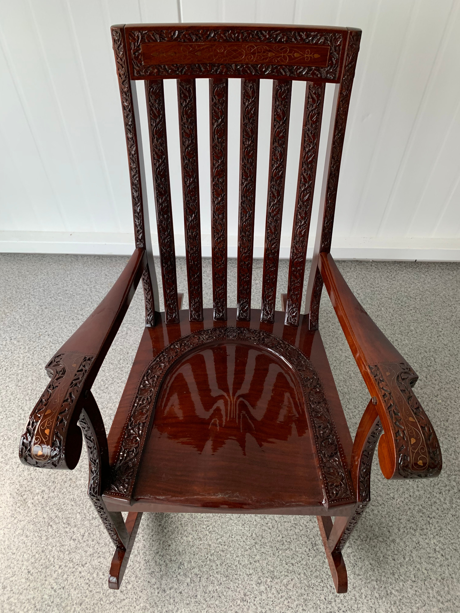 Bogato zdobiony, ciemnobrązowy fotel z rzeźbieniami, widok z góry. Ornamenty na oparciu, podłokietnikach i siedzisku. Polerowane drewno z widocznymi refleksami światła.