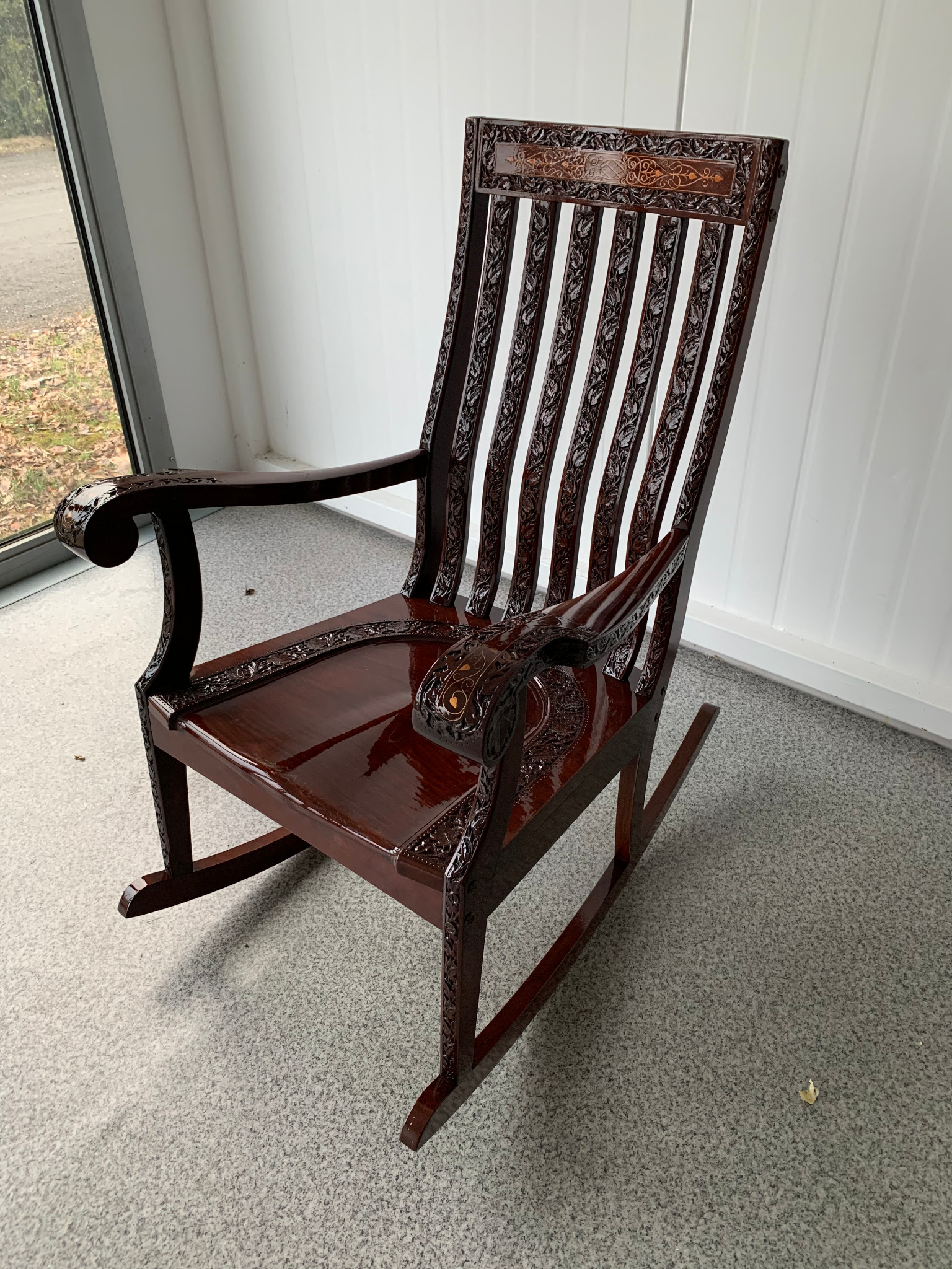 Ciemnobrązowy, lakierowany fotel bujany z rzeźbionymi detalami w stylu retro, ustawiony w jasnym pomieszczeniu z oknem.