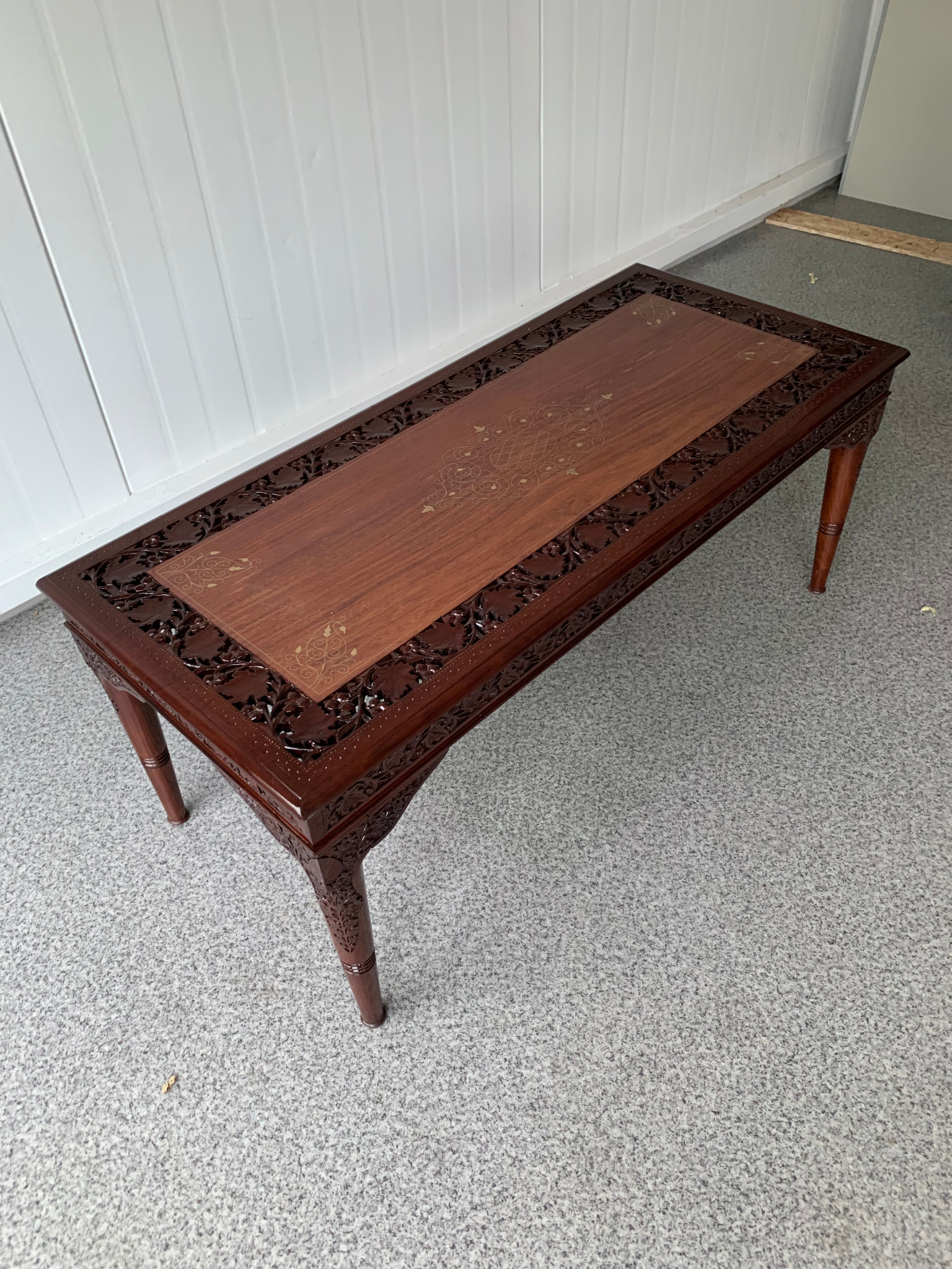 Ręcznie rzeźbiony stolik kawowy z ciemnego drewna, z jasnymi intarsjami na blacie, ustawiony na szarym dywanie i tle z białych paneli ściennych.