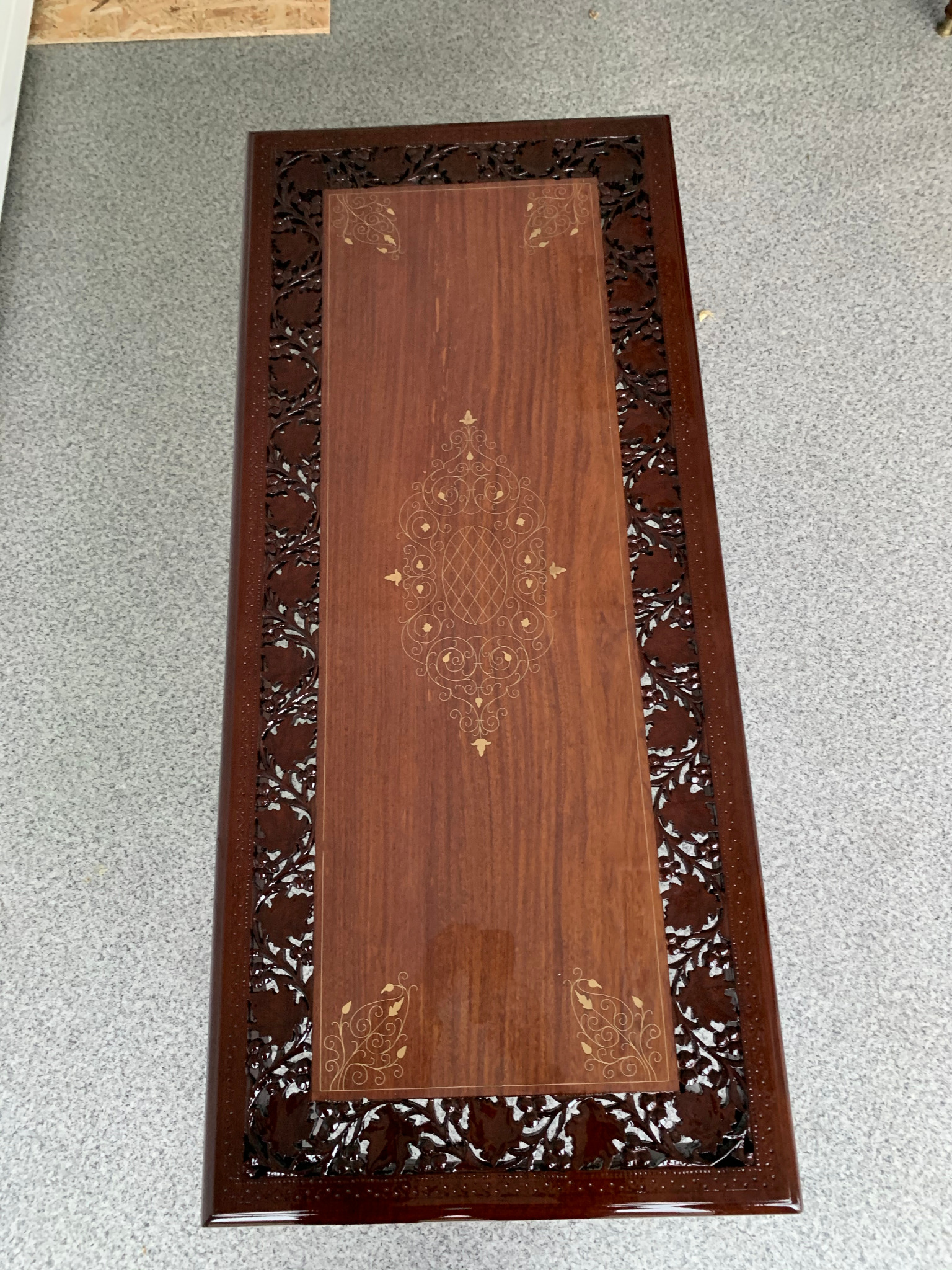 Lakierowany stolik kawowy z ciemnego drewna z rzeźbionymi krawędziami i złoconymi zdobieniami na blacie, widziany z góry na tle szarej podłogi.