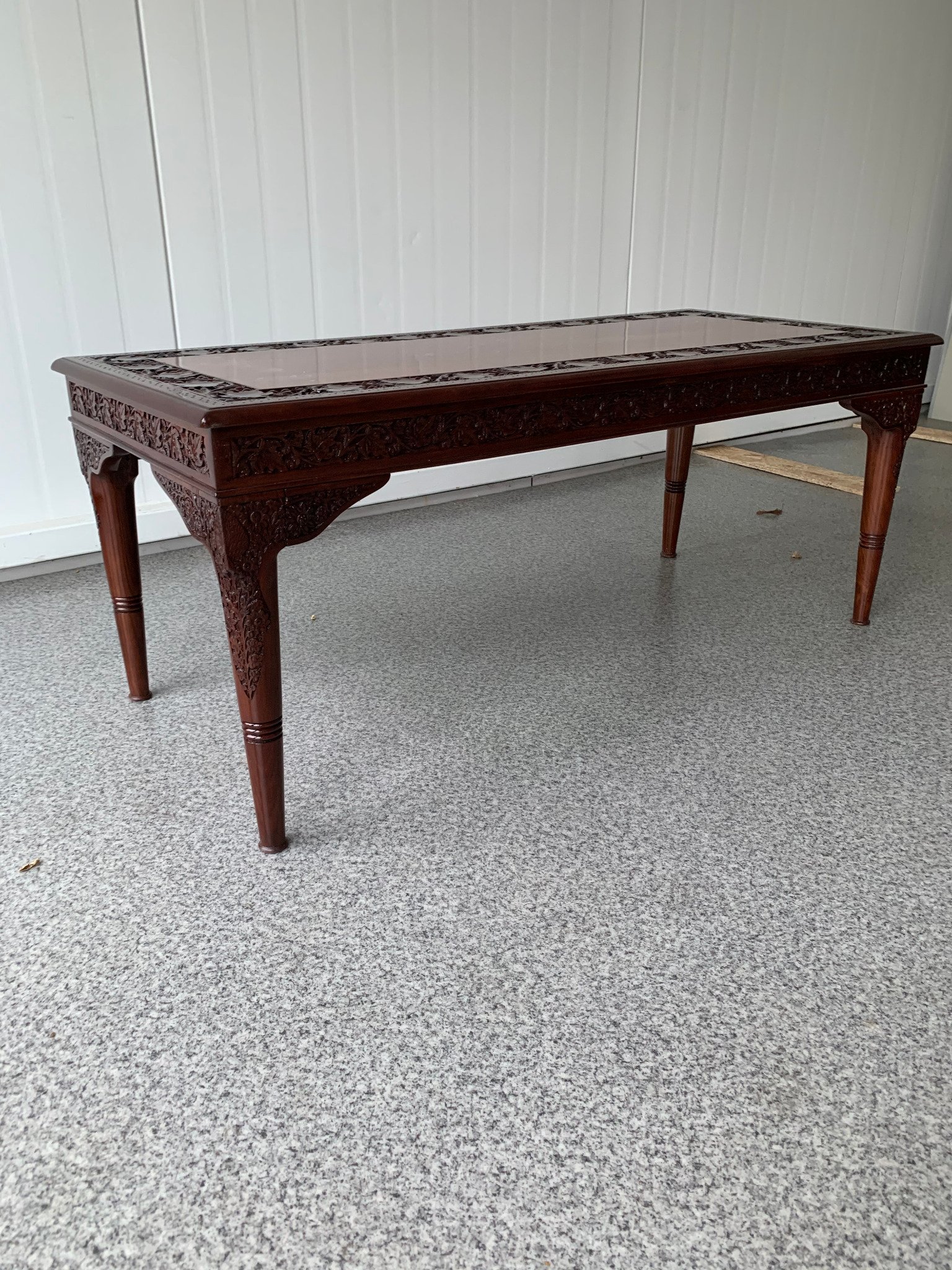 Ręcznie rzeźbiony, mahoniowy stolik kawowy z szklanym blatem na tle białej ściany. Detale rzeźbienia na blacie i nogach.