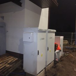 Krystian Russek - Instalacja elektryczna zewnętrzna: trzy szare szafy elektryczne na betonowych podstawach, pracownik w pomarańczowej kamizelce wykonuje podłączenie. Widoczne palety i wykop.