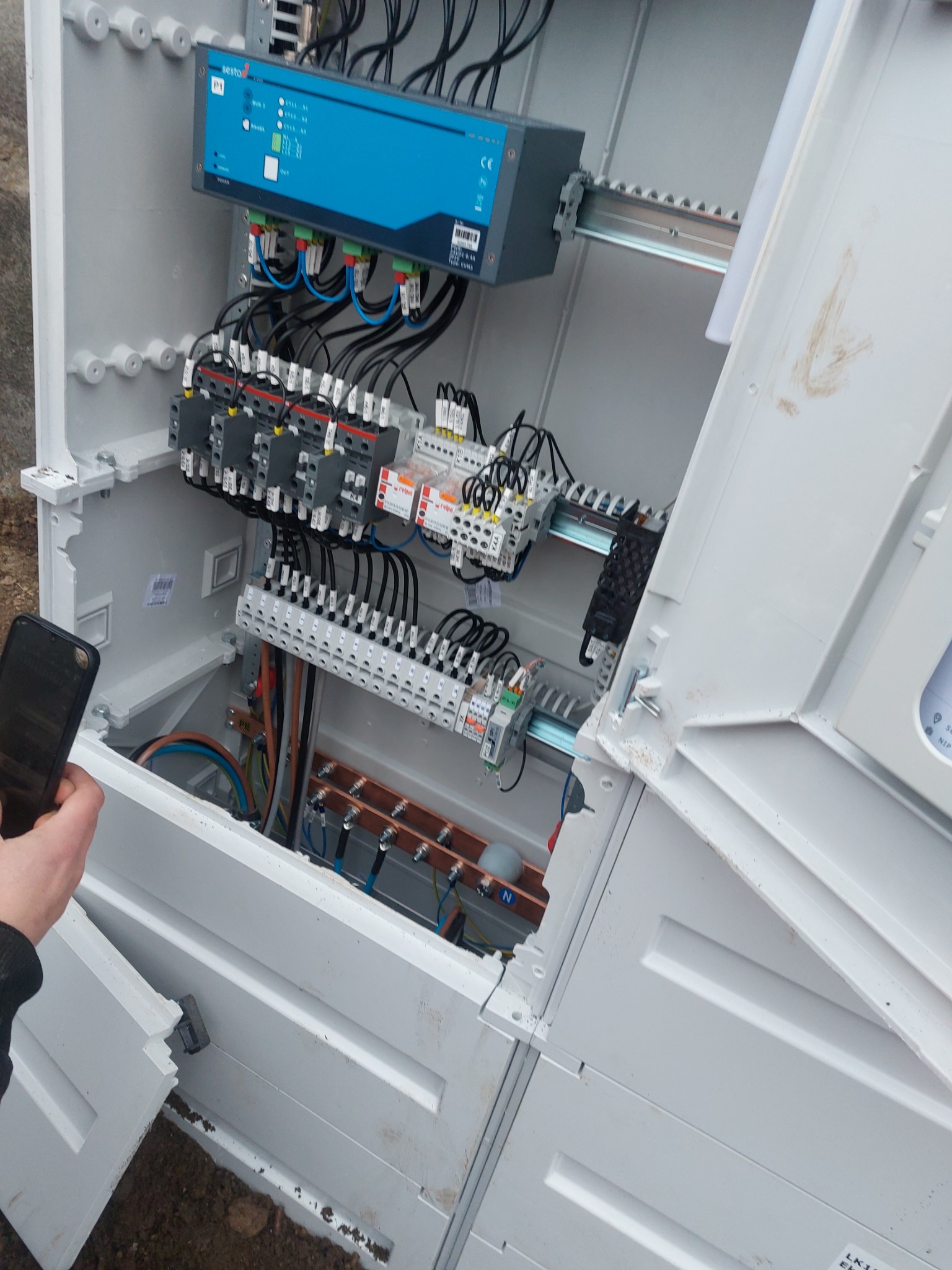 Otwarta skrzynka elektryczna z zainstalowanymi elementami, widoczne kable, bezpieczniki i listwy. Osoba trzyma telefon.