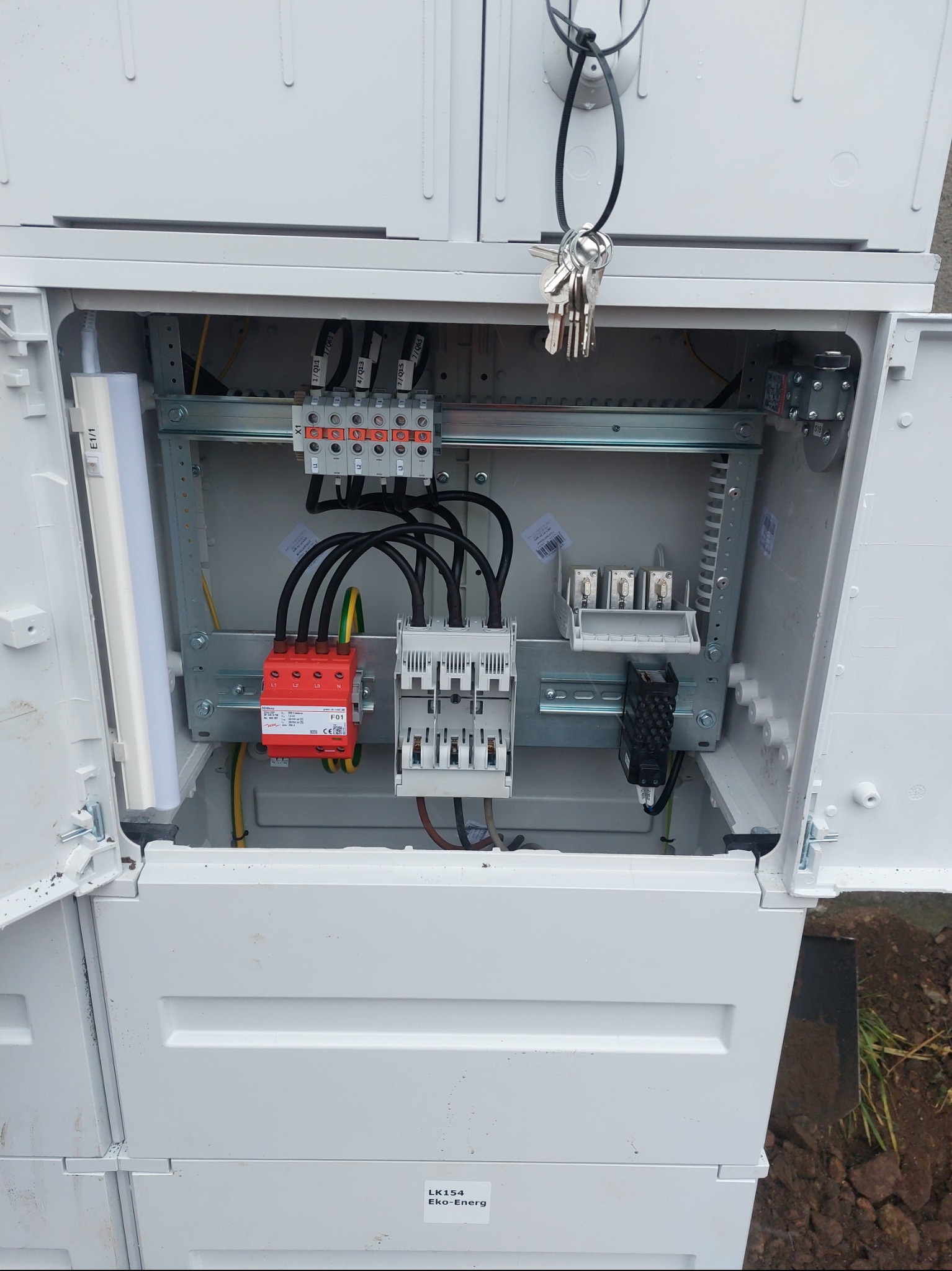 Otwarta, biała skrzynka elektryczna z zainstalowanymi elementami: bezpieczniki, przewody, listwy zaciskowe. Widoczne klucze na górze skrzynki.