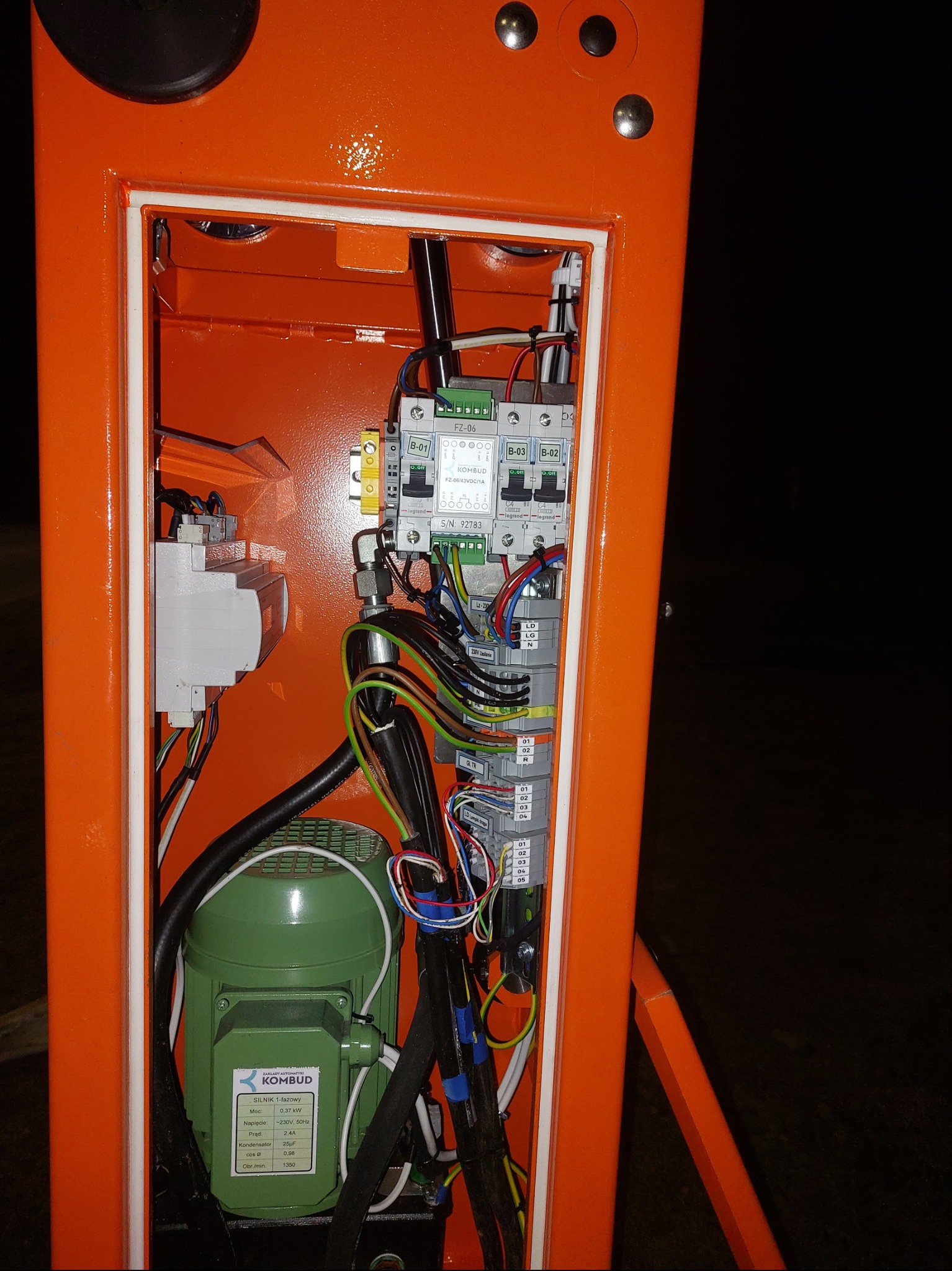 Otwarta, pomarańczowa skrzynka elektryczna z widocznymi przewodami, bezpiecznikami i zielonym silnikiem KOMBUD. Widoczne oznaczenia i etykiety na elementach.
