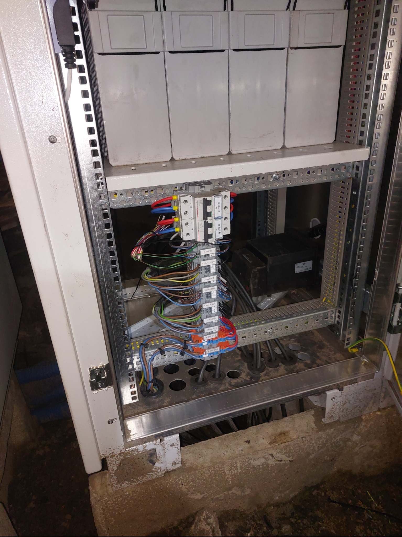 Rozdzielnia elektryczna w metalowej obudowie z widocznymi przewodami, bezpiecznikami i elementami montażowymi. Widoczne kable w różnych kolorach.