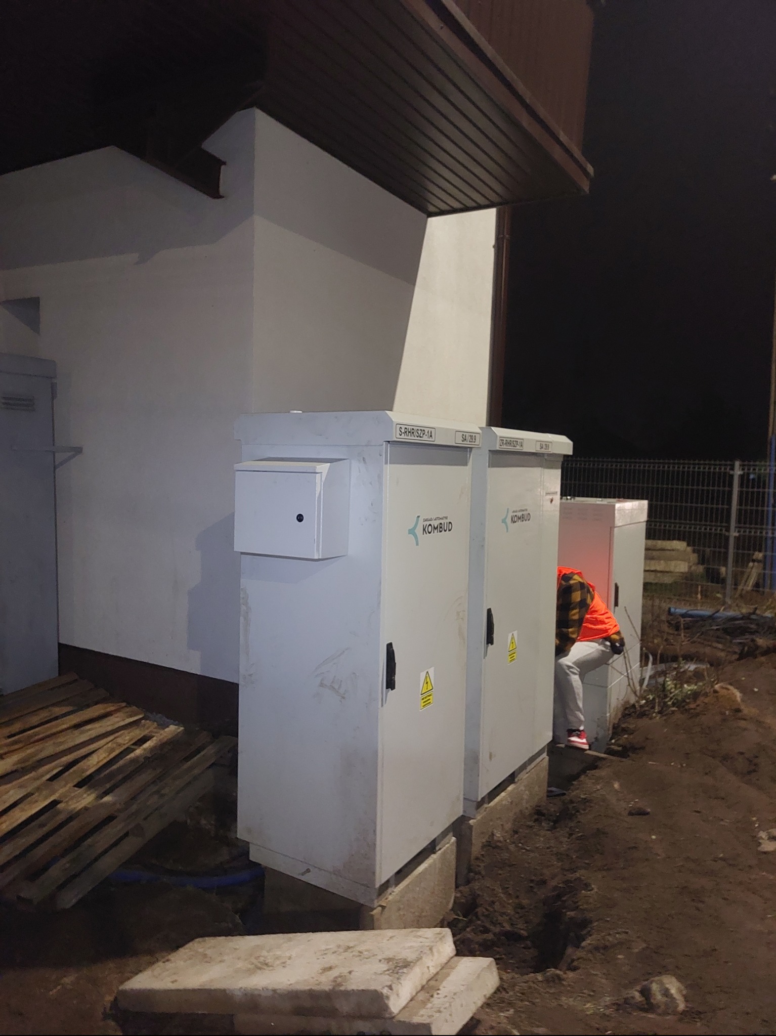 Instalacja elektryczna zewnętrzna: trzy szare szafy elektryczne na betonowych podstawach, pracownik w pomarańczowej kamizelce wykonuje podłączenie. Widoczne palety i wykop.