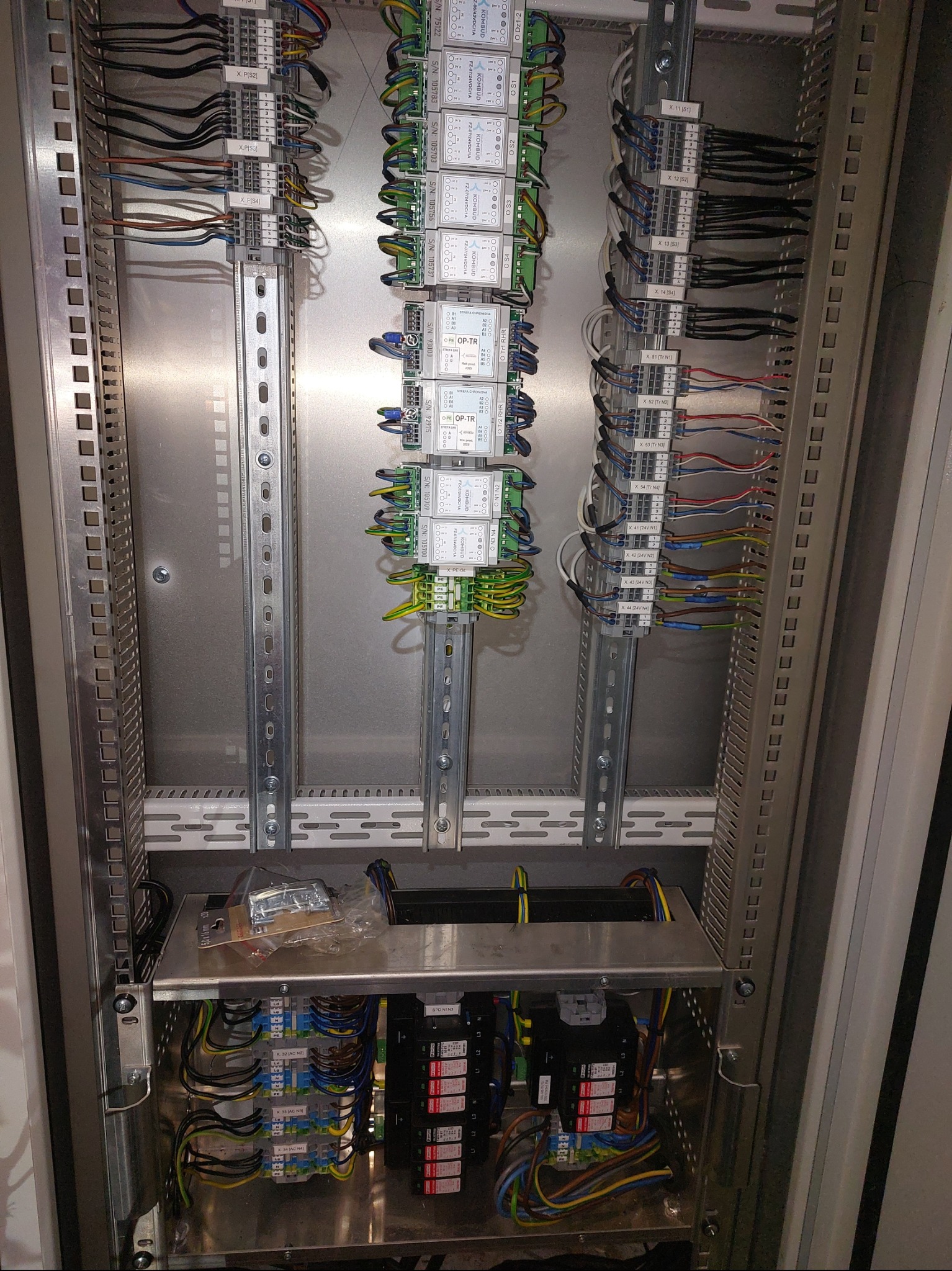 Wnętrze szafy sterowniczej z modułami, przewodami i listwami zaciskowymi. Widoczne elementy automatyki przemysłowej i okablowanie. Precyzyjny montaż w metalowej obudowie.