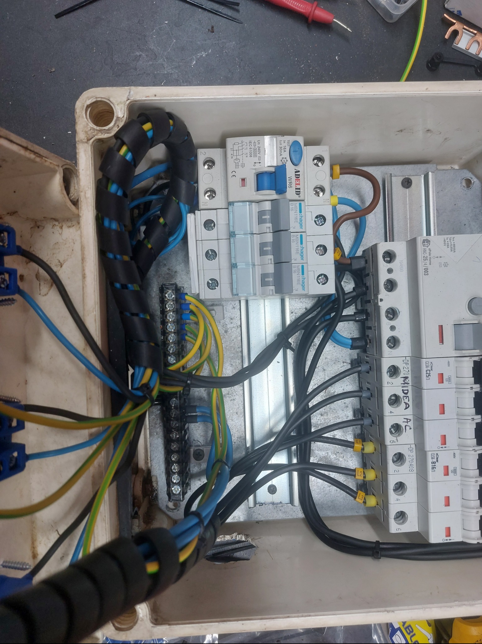 Otwarta skrzynka elektryczna z zabezpieczeniami, przewodami w peszlu i listwą zaciskową. Widoczne bezpieczniki i oznaczenia. Instalacja w trakcie prac.