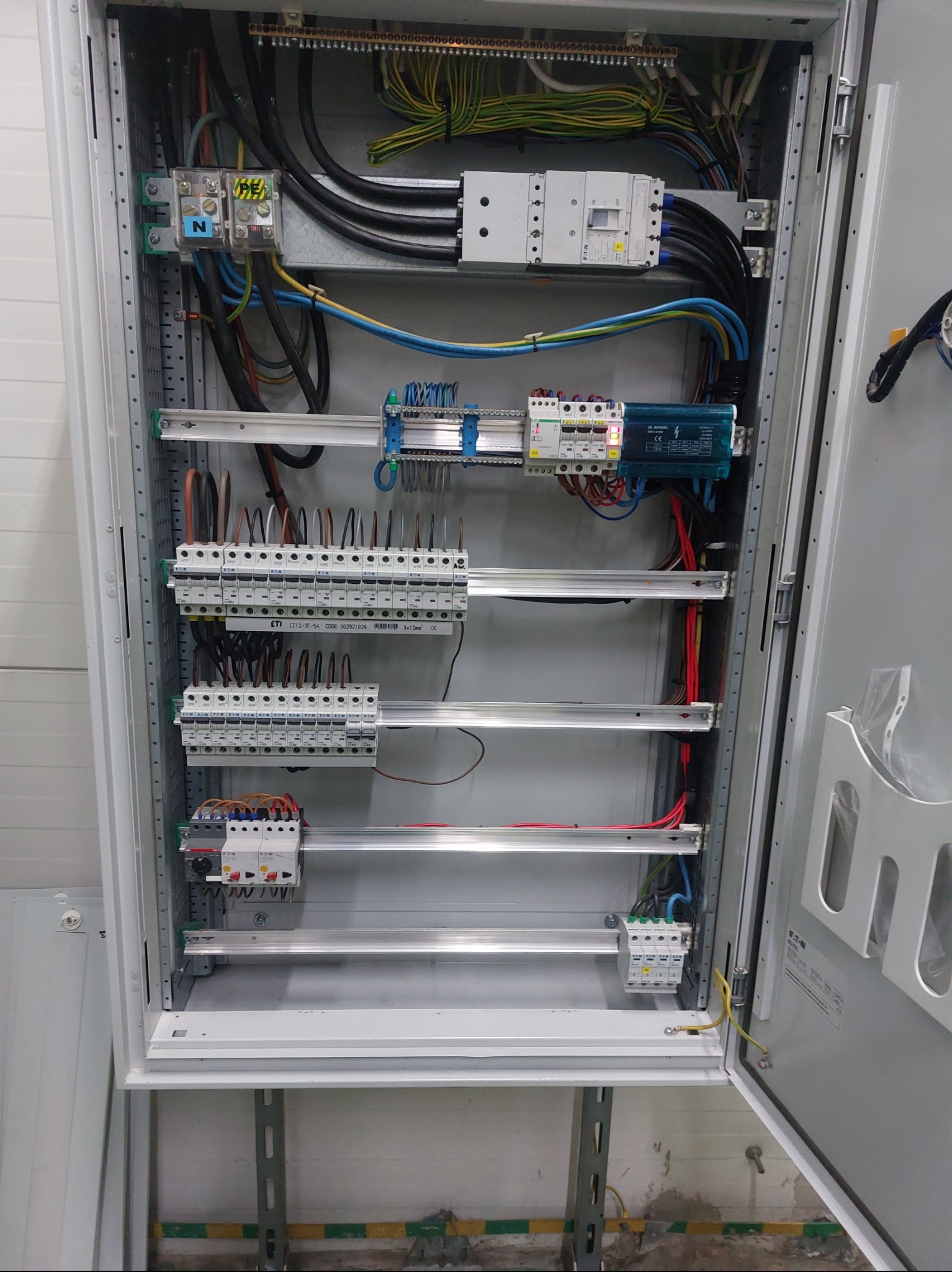 Otwarta szafa elektryczna z widocznymi bezpiecznikami, przewodami i elementami montażowymi. Widoczne kable w różnych kolorach i oznaczenia na urządzeniach.