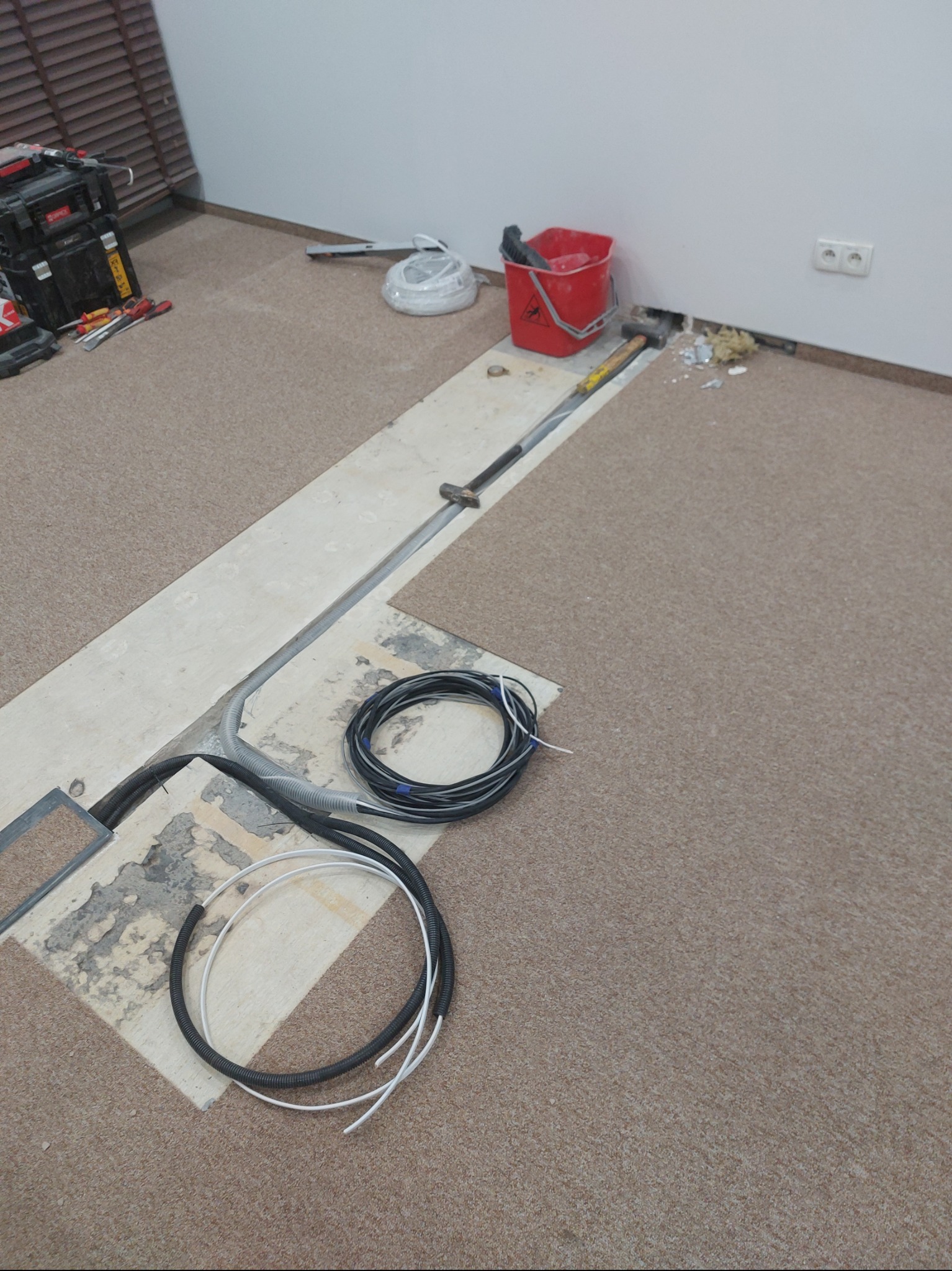 Kable w podłodze podczas prac remontowych. Widoczne narzędzia, wiadro i gniazdko elektryczne. Fragment podłogi z wykładziną. Prace w toku.