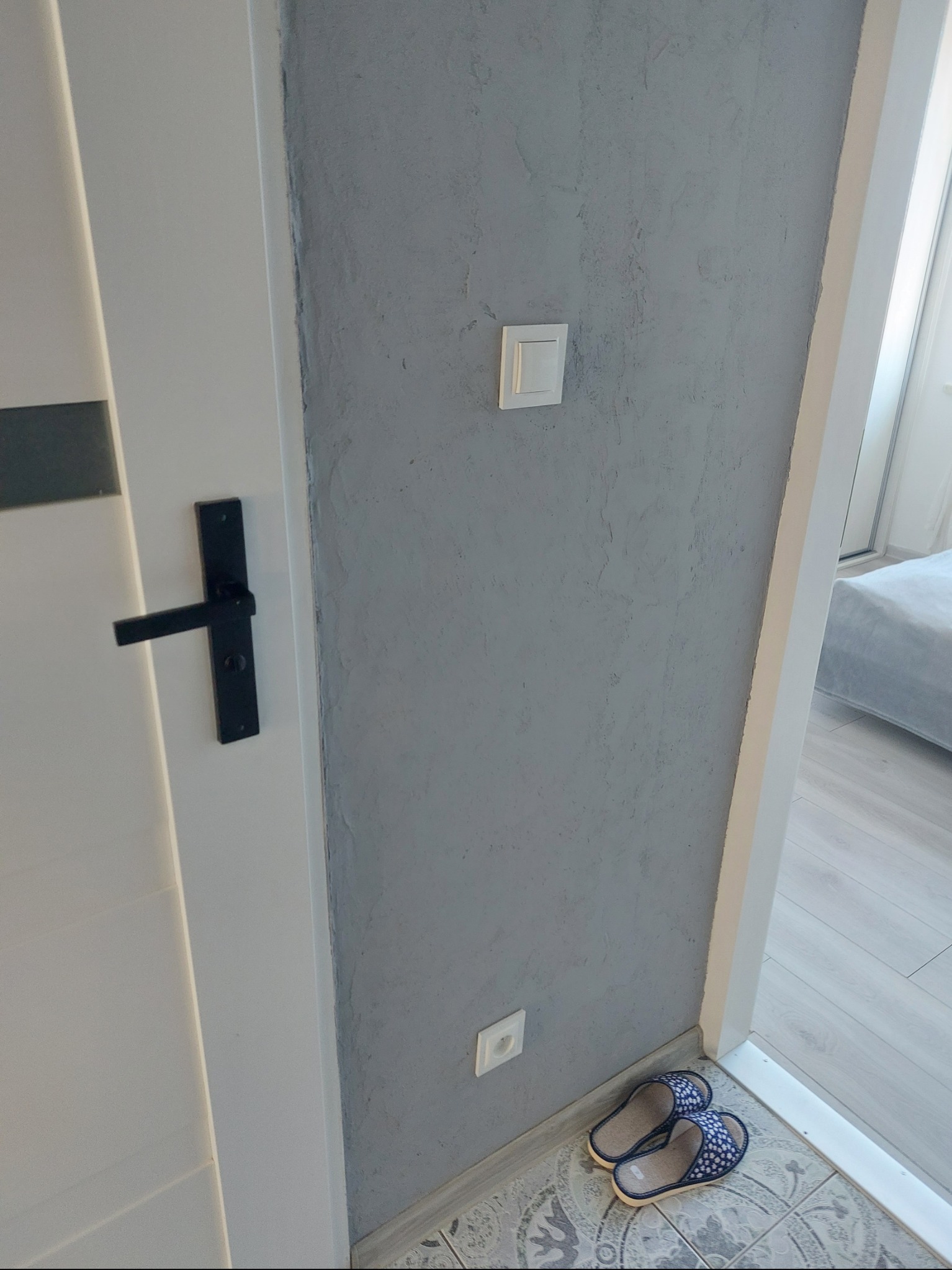 Szara ściana z fakturą, biały włącznik i gniazdko. Na podłodze wzorzyste płytki i kapcie. Widok na pokój z łóżkiem przez otwarte drzwi.