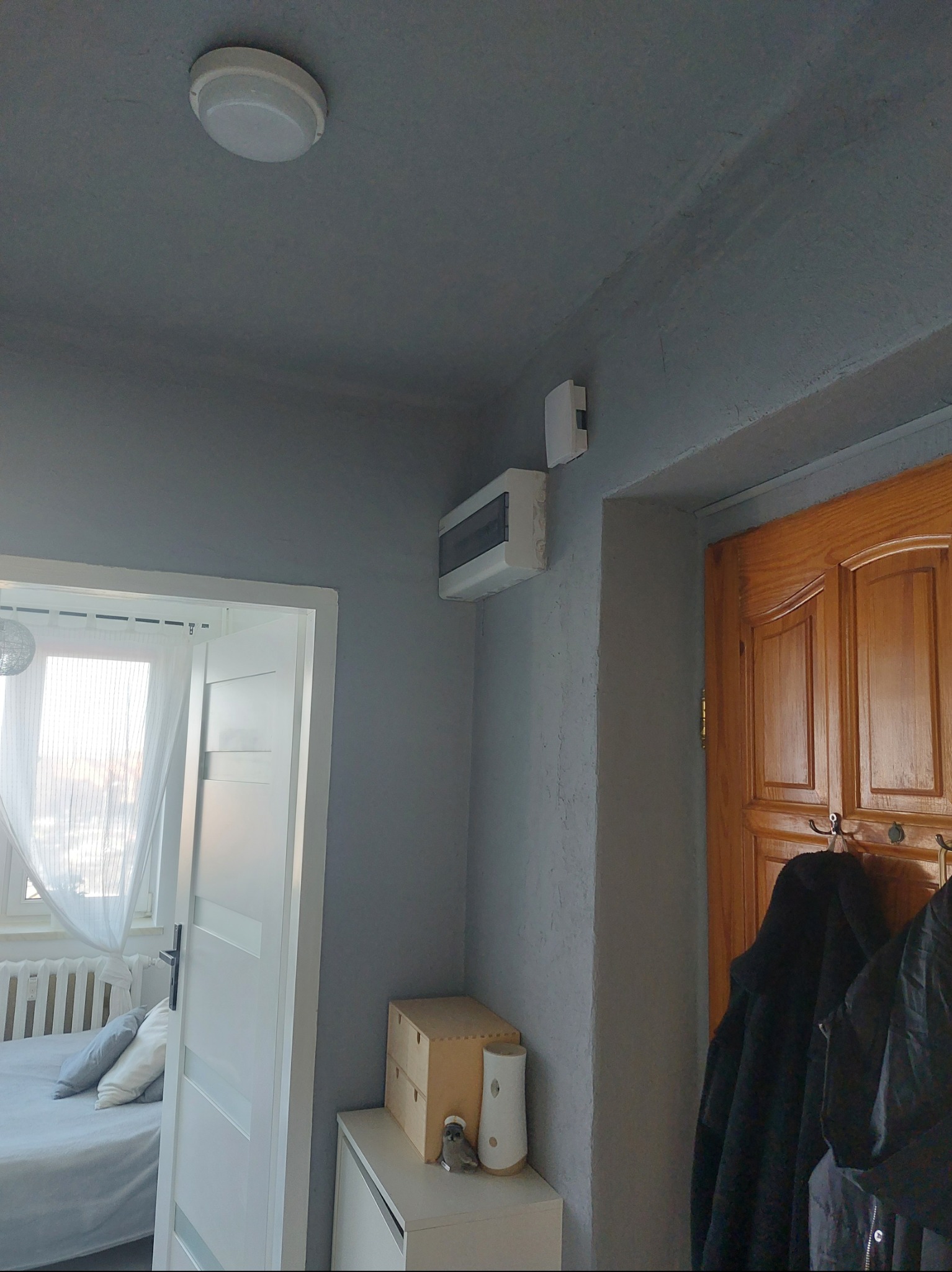 Szara ściana z zamontowaną białą skrzynką elektryczną, widoczny fragment drewnianych drzwi i białe drzwi do pokoju z łóżkiem. Na suficie okrągła lampa.
