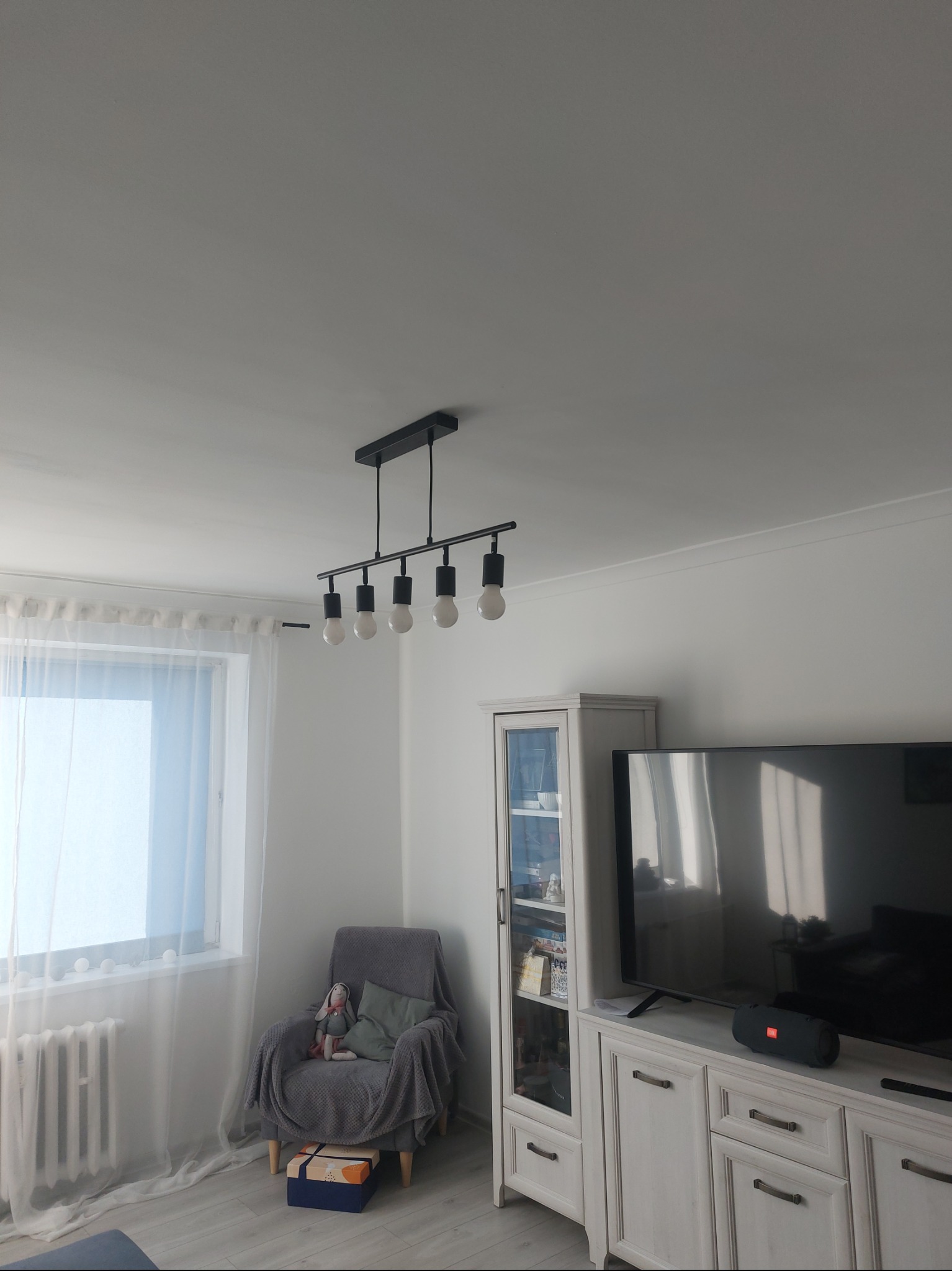 Nowoczesna lampa sufitowa z pięcioma żarówkami w minimalistycznym salonie z szarym fotelem i telewizorem. Jasne wnętrze z subtelnym oświetleniem.