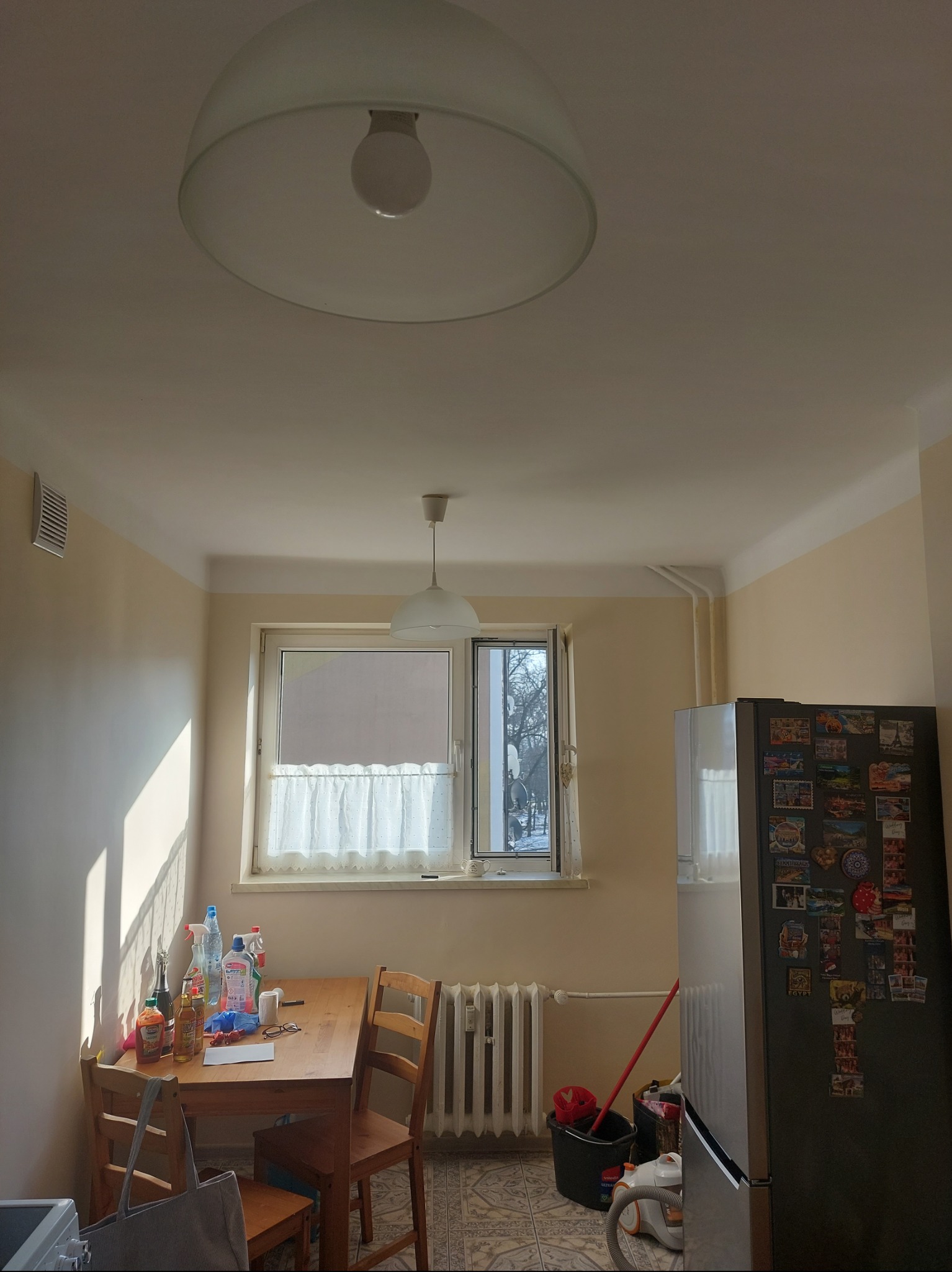 Jasne wnętrze kuchni z drewnianym stołem, krzesłami i lampami sufitowymi. Widoczne okno z firanką, lodówka z magnesami i akcesoria do sprzątania.